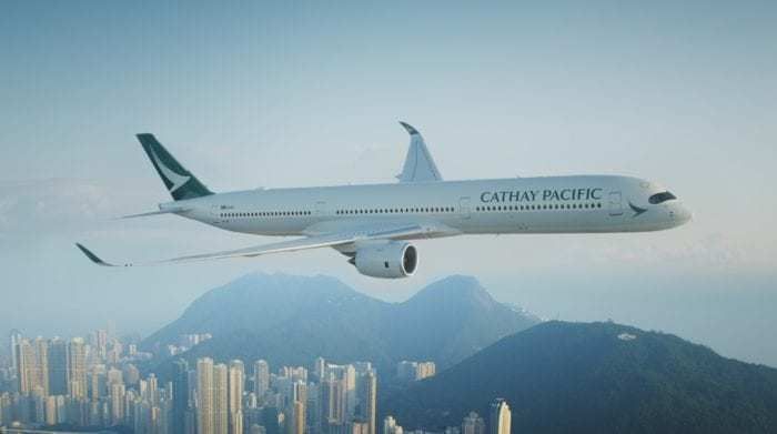 A Cathay Pacific Airbus A350-1000 flying low, with the Hong Kong city skyline in the background.