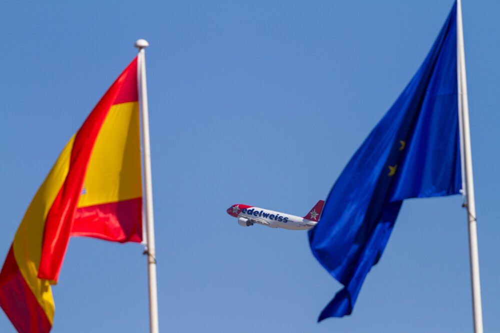 An Edelweiss Airbus A320 flying in the sky behind an EU Flag.