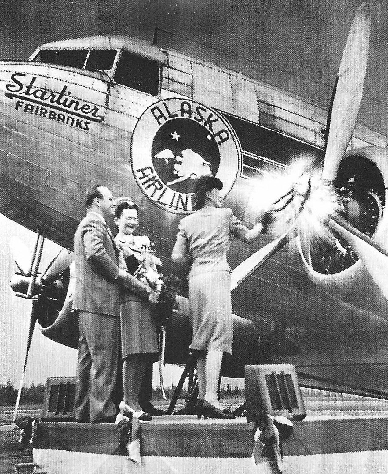 Starliner Fairbanks Christening With A Man & Two Women in 1940s Formal Clothes Standing While One Woman Breaks Champagne on the DC-3's Left Propeller Hub