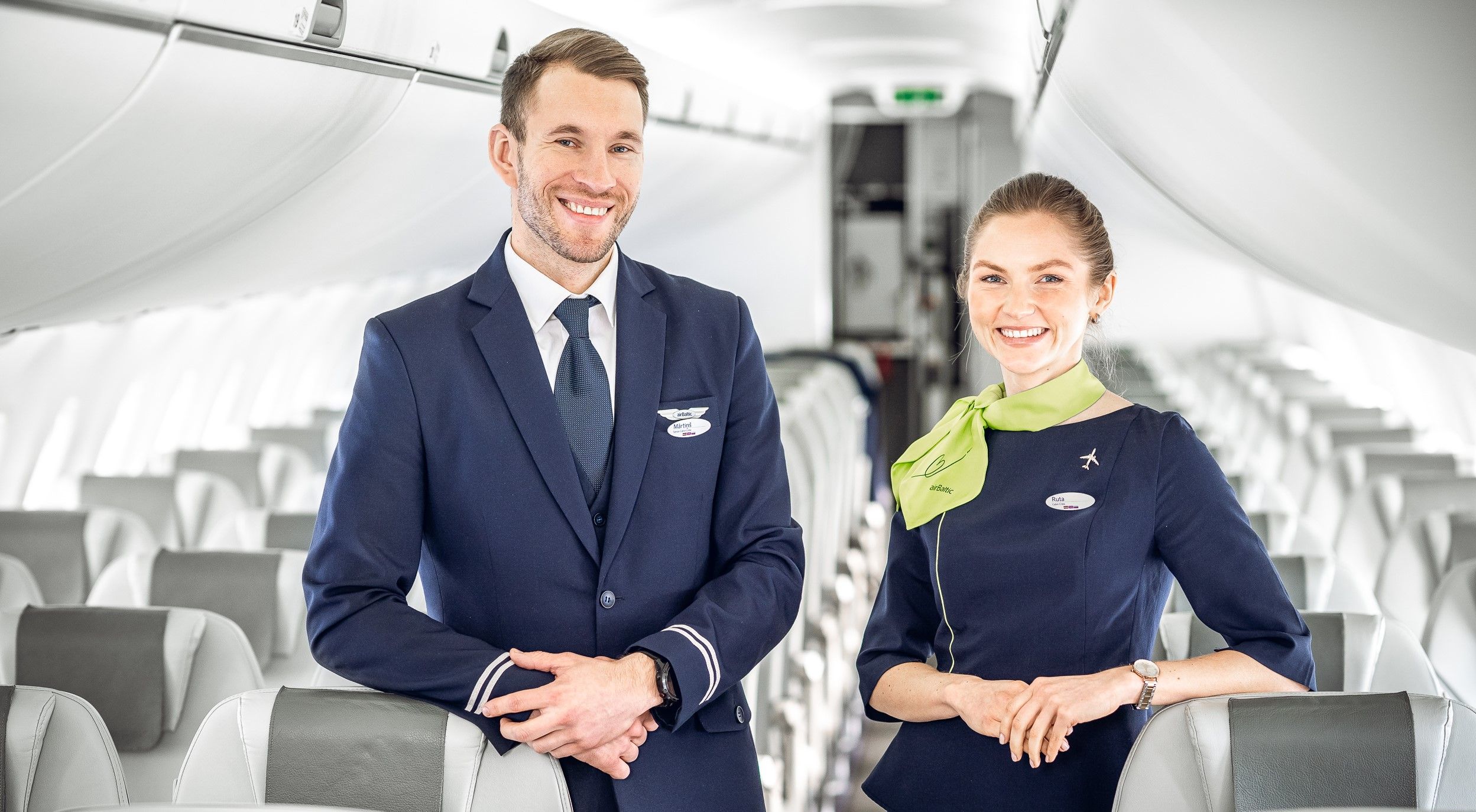 Two airBaltic cabin crew members standing in the cabin of a narrowbody aircraft.