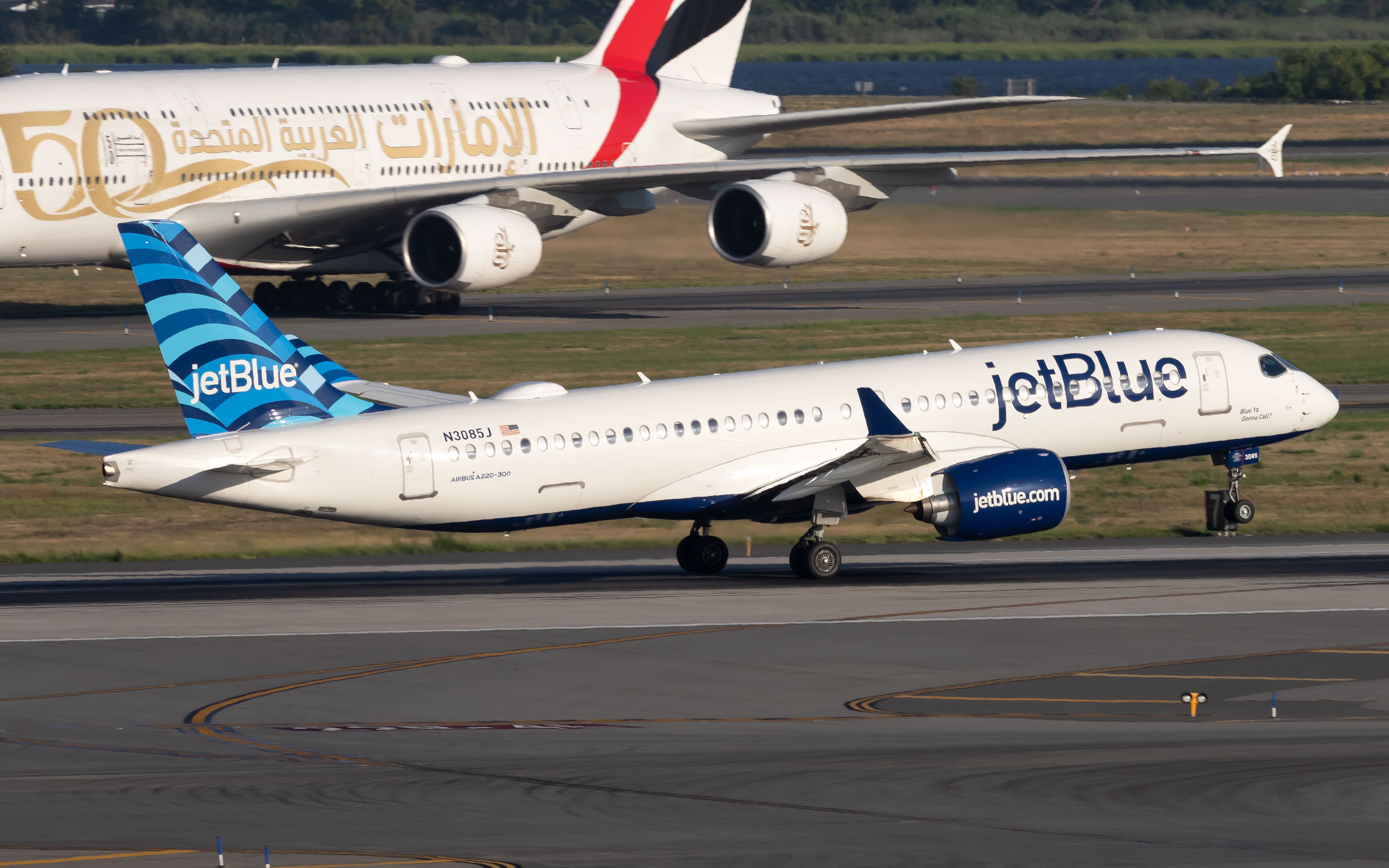 A jetBlue Airbus A220-300 about to takeoff, while an Emirates A380 waits in the background.
