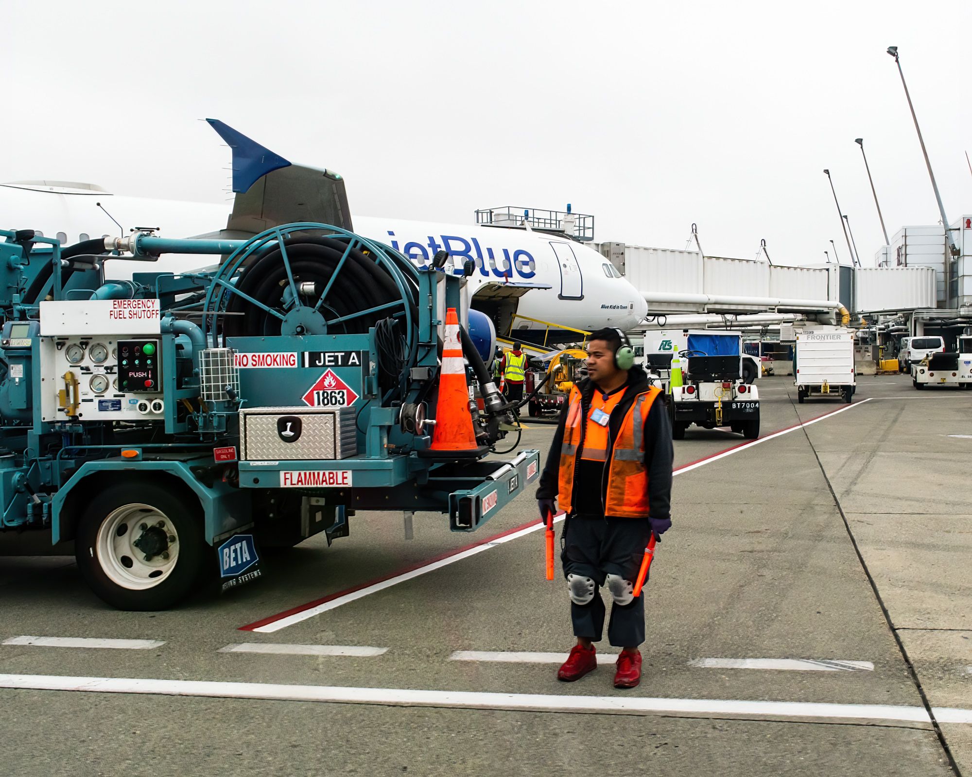 A ground crew worker standing on an airport apron next to a refueling truck and a JetBlue Airbus A321.