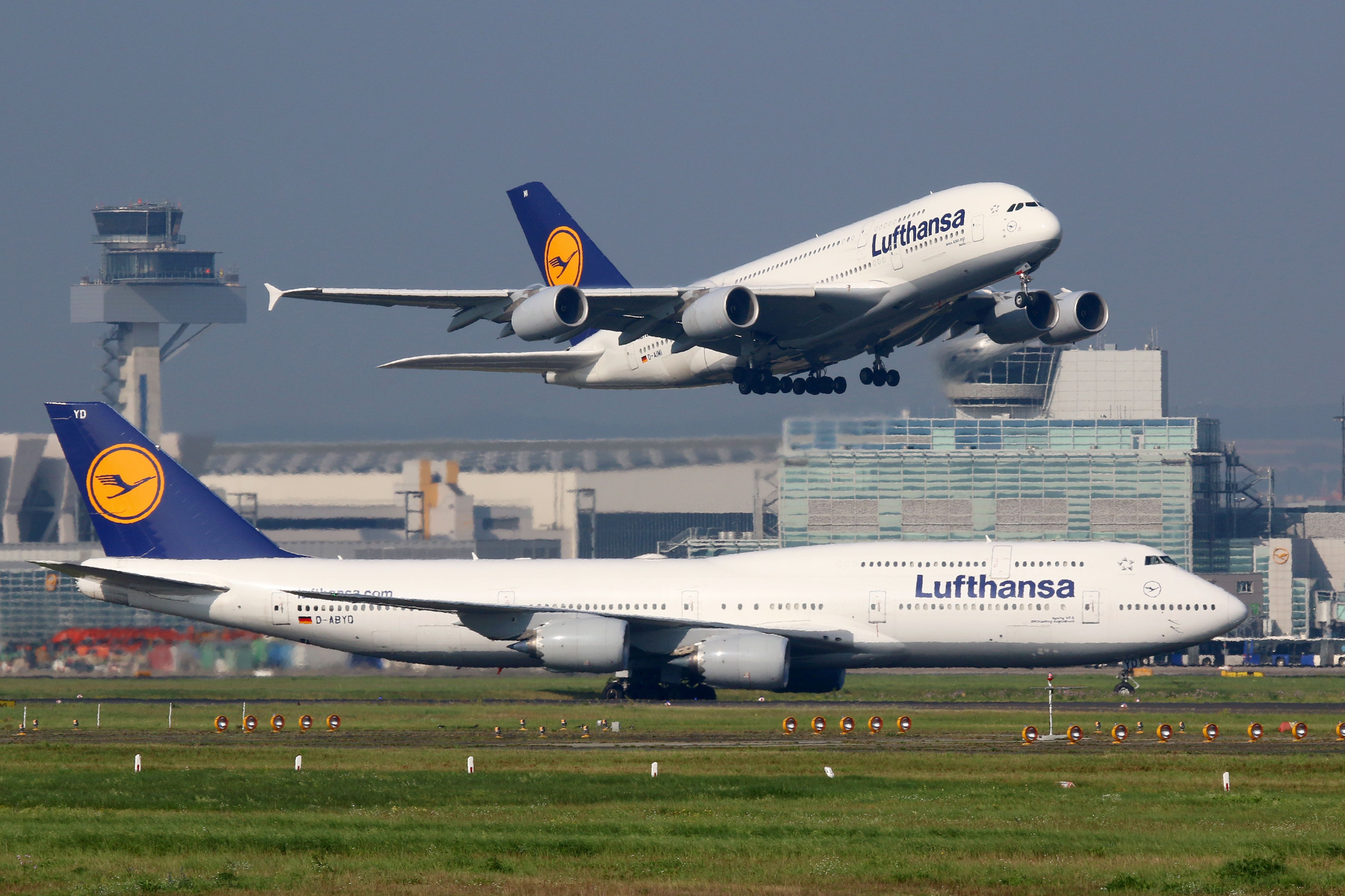 A Lufthansa Airbus A380 taking off while a Lufthansa Boeing 747 sits on a taxiway.