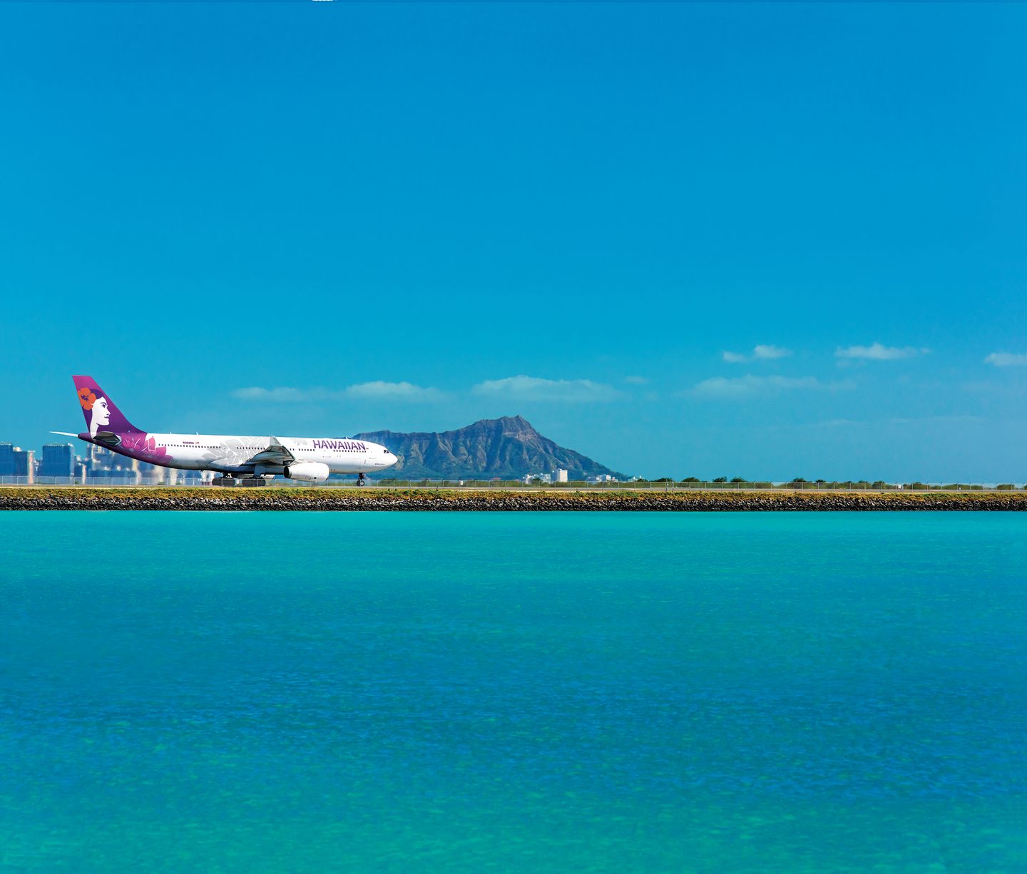 White Airbus 330 aircraft with pink and purple Hawaiian Airlines branding on a runway with a mountain in the background and blue ocean in the foreground