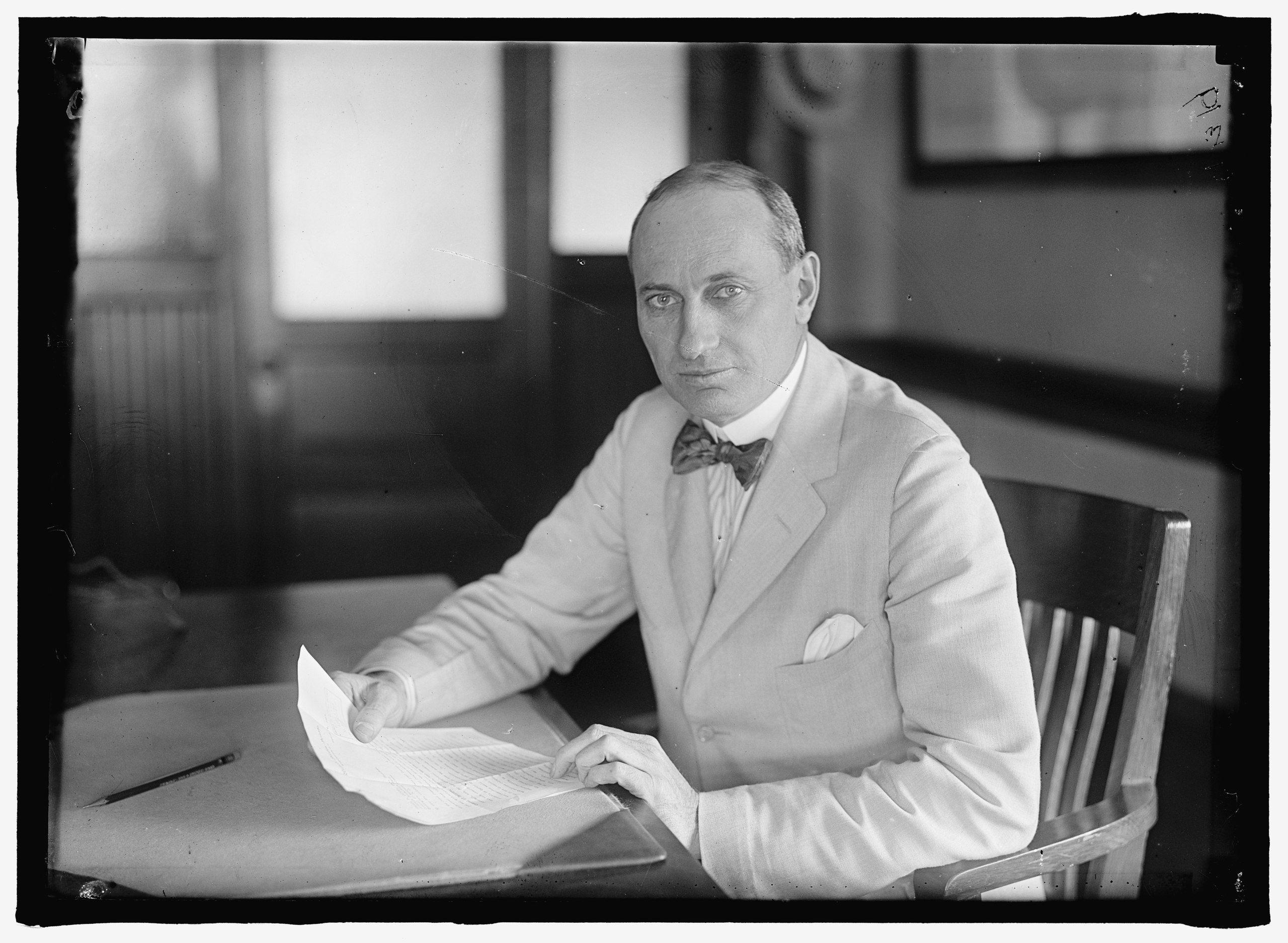 A man wearing a blazer and bow tie sitting a desk holding documents