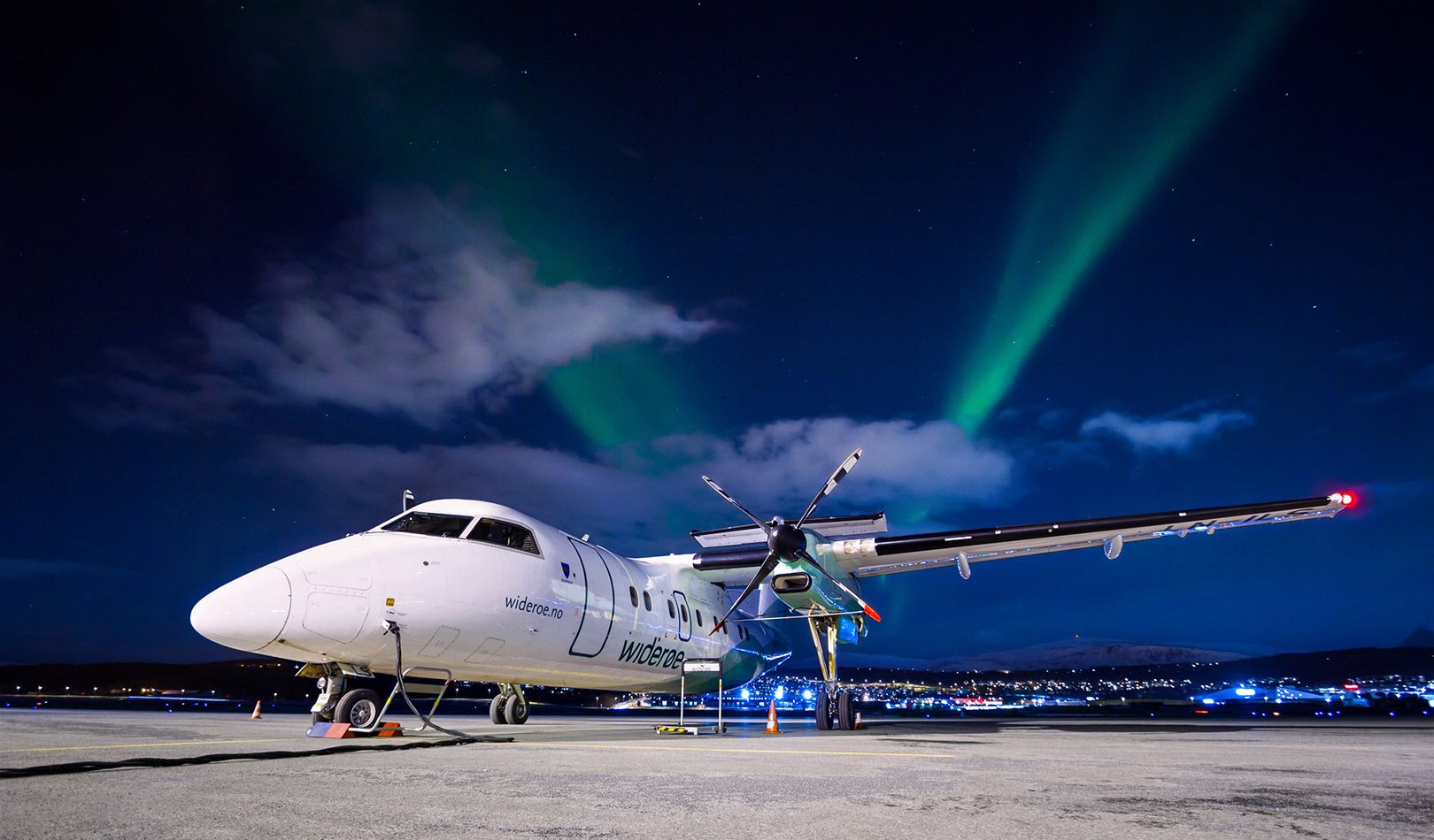 A Wideroe aircraft parked. It is night in the winter and a few northern lights can be seen in the back.