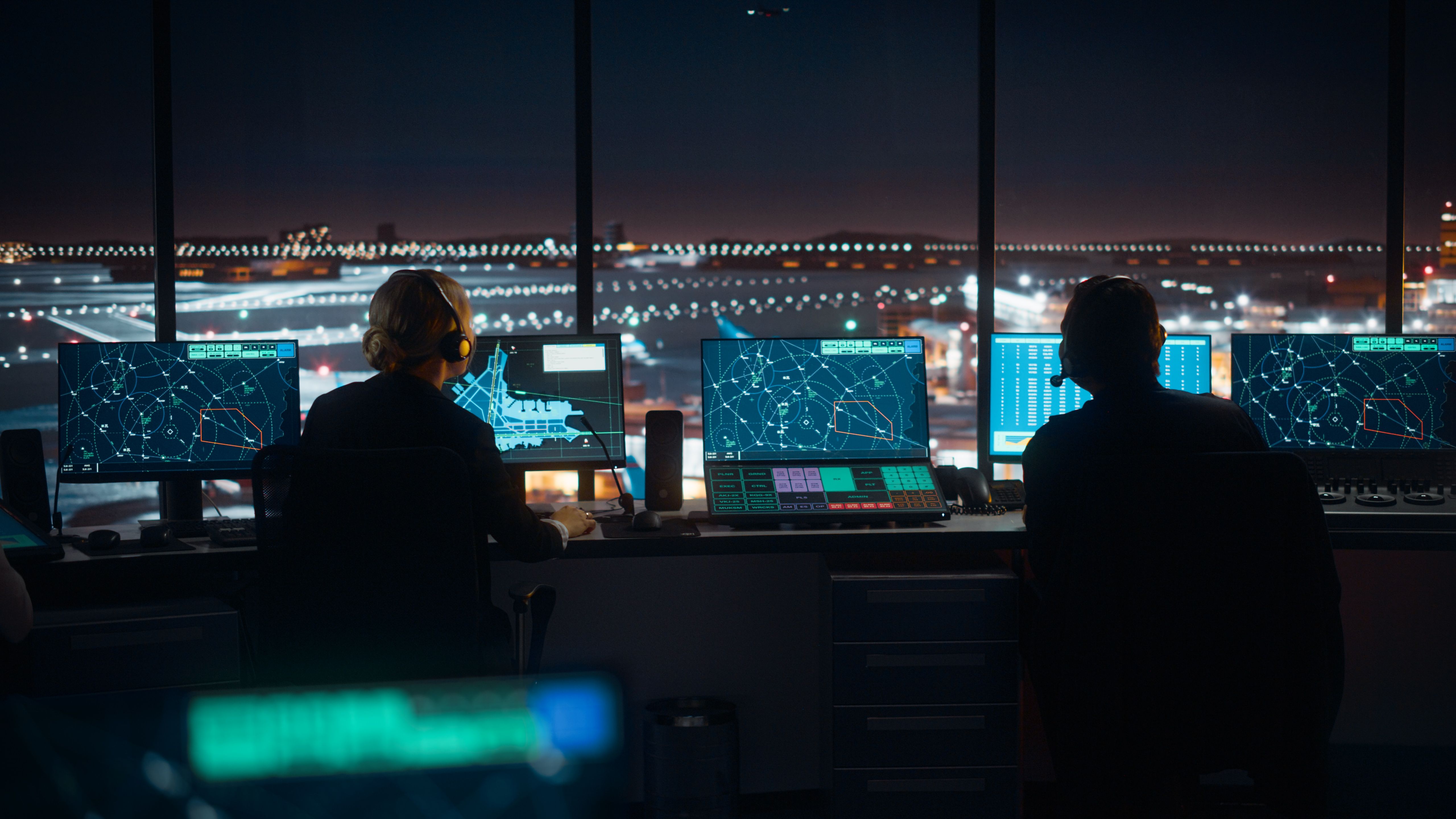 Two workers sitting behind Air Traffic Control Displays in a tower overlooking the airport apron.