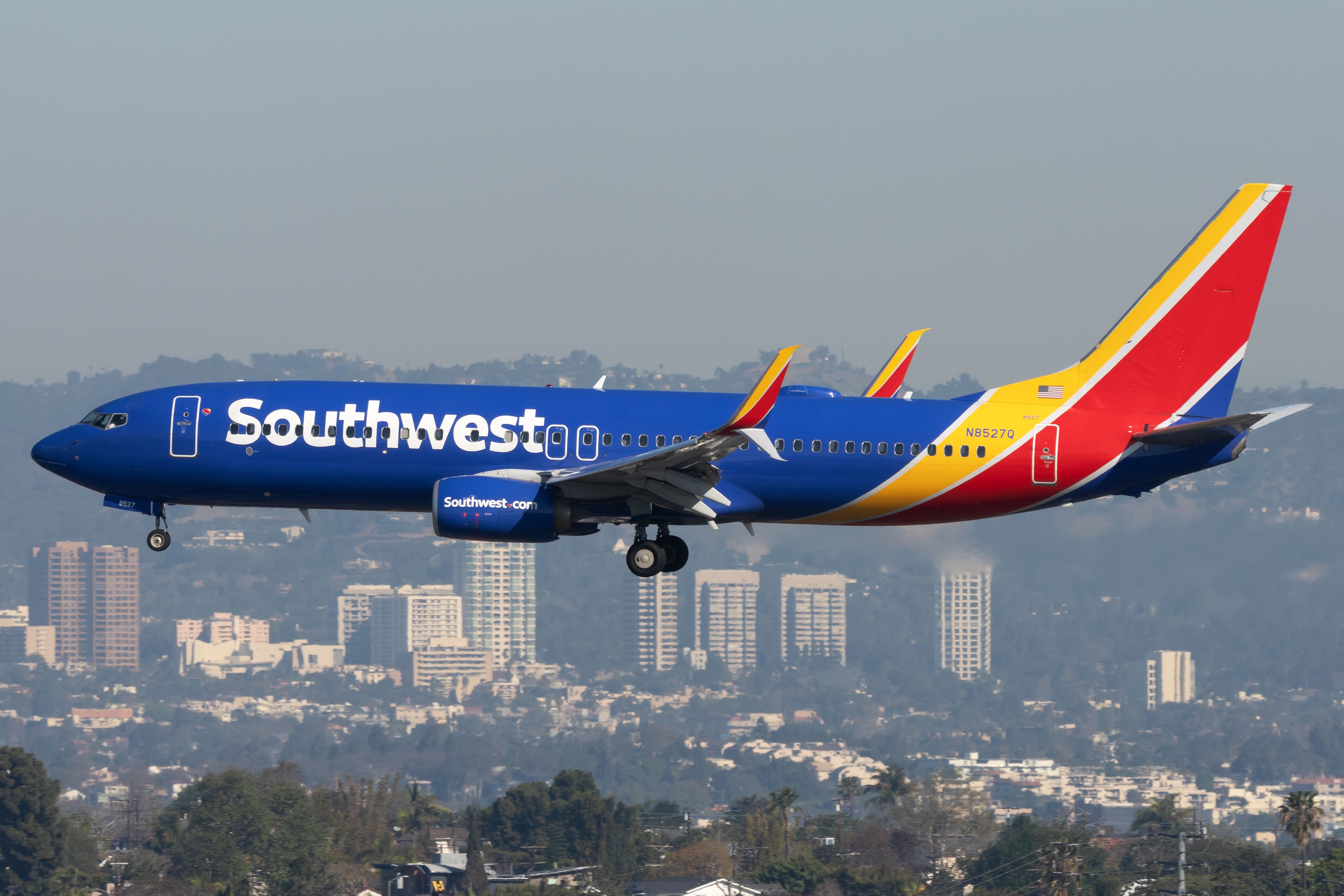 A blue Southwest Airline Boeing 737 with white lettering and a red and yellow tail flying near an urban area with tall buildings in the background