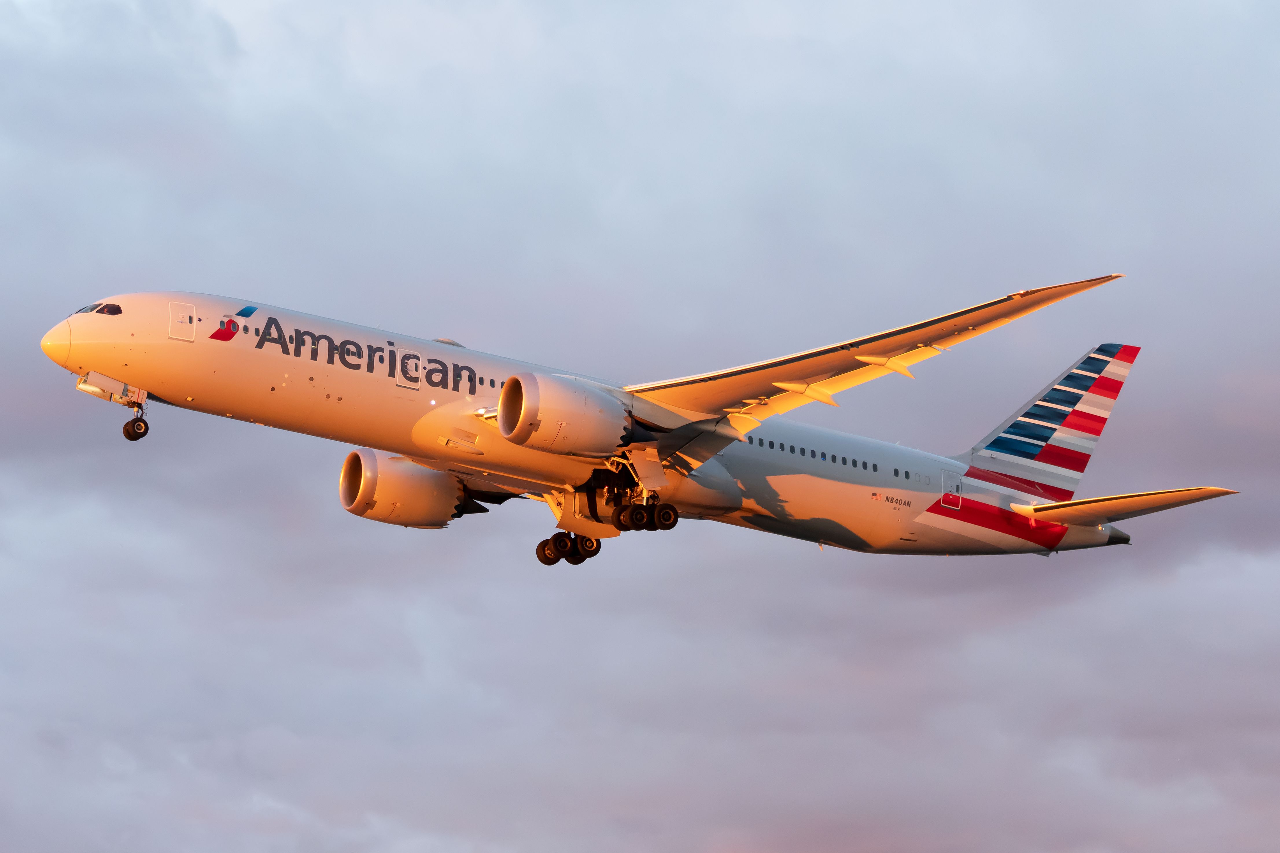 White Boeing 787 jetliner with red and blue stripes on the tail and American in blue lettering on the side flying in cloudy sky