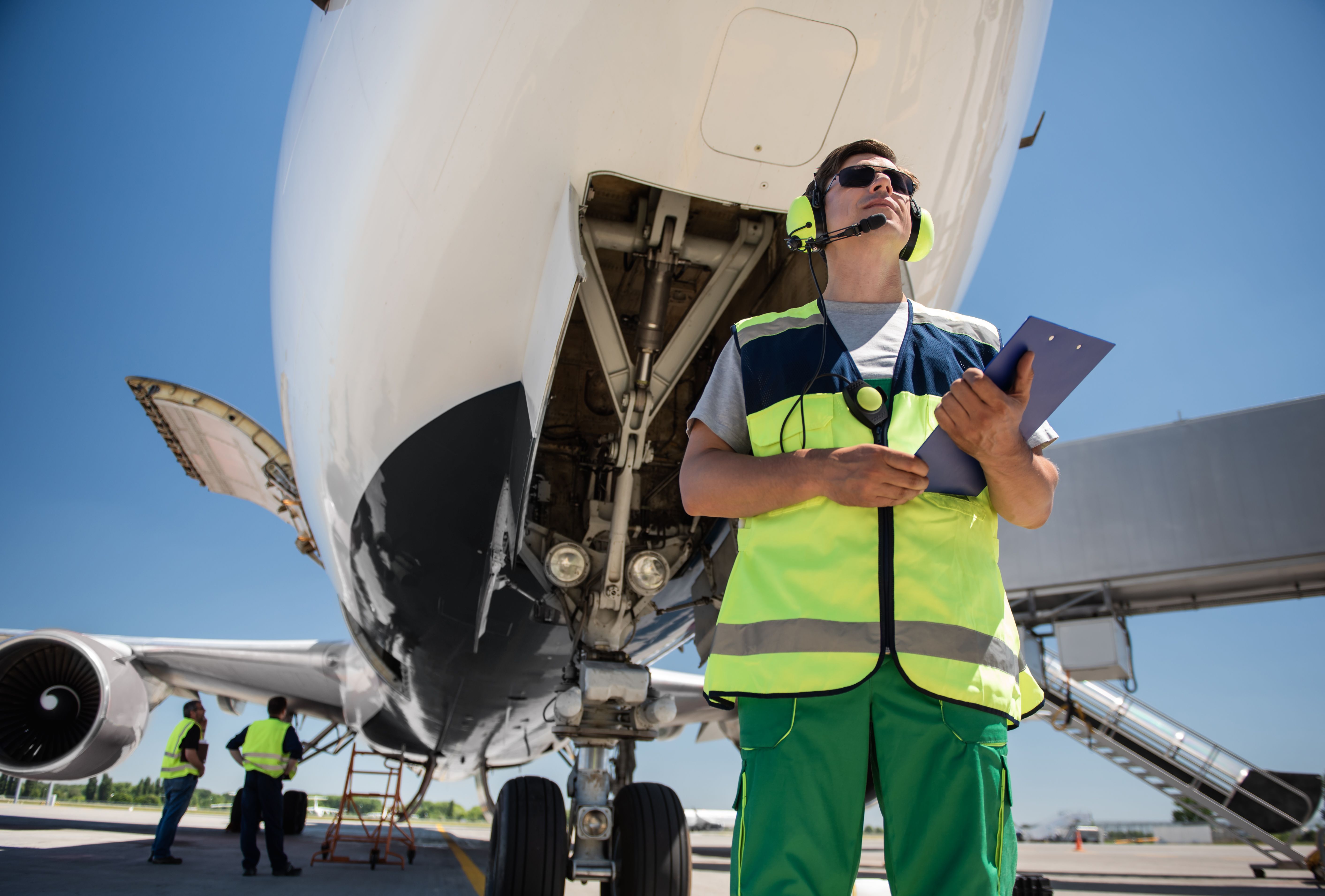 Preparation for the flight. Low angle portrait of man in sunglasses holding documents and posing near aircraft with open door. Ground crewmembers standing behind 