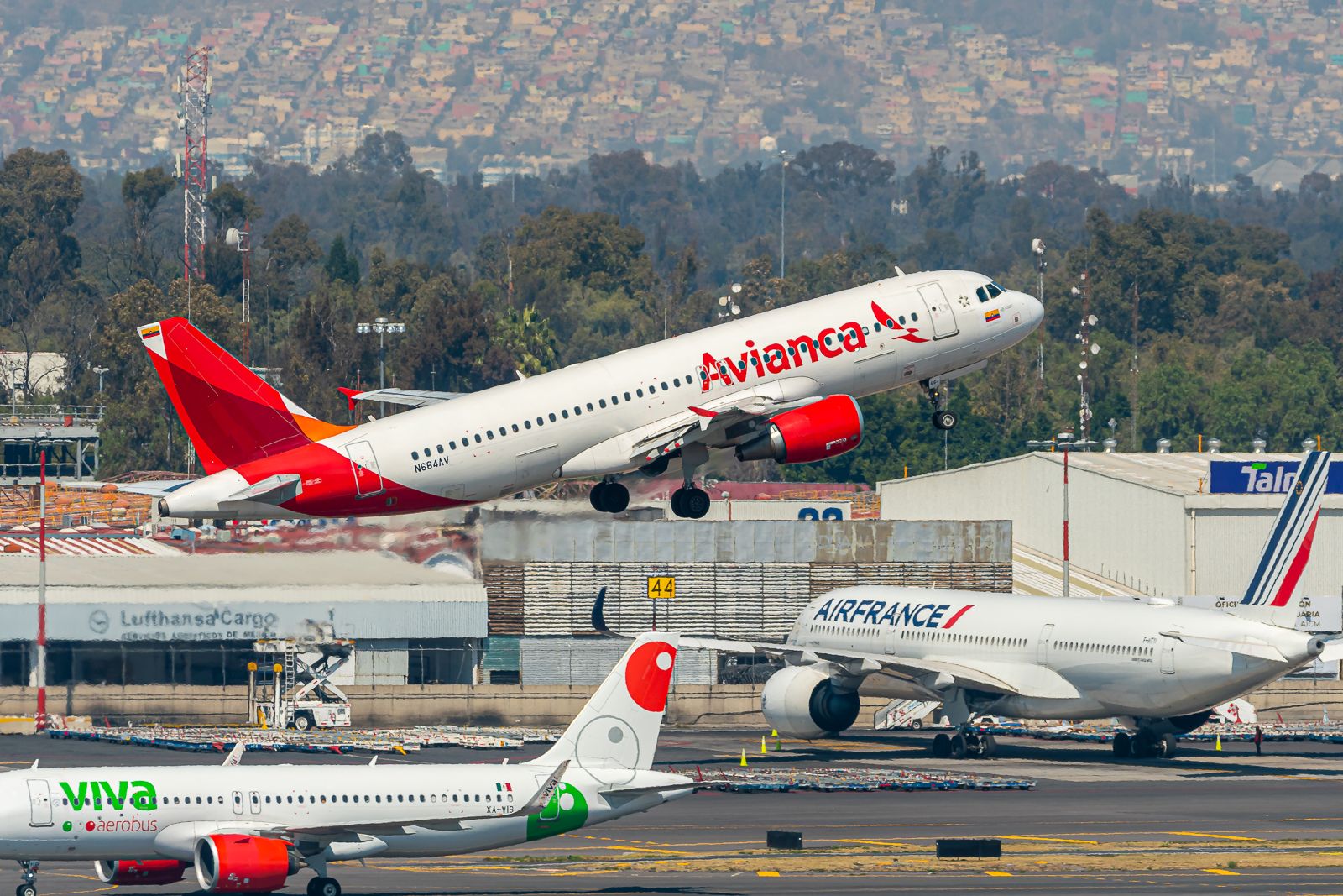 An Avianca aircraft departing from Mexico City. In the back, a Viva Aerobus aircraft can be seen taxiing and an Air France aircraft can be seen parked. 