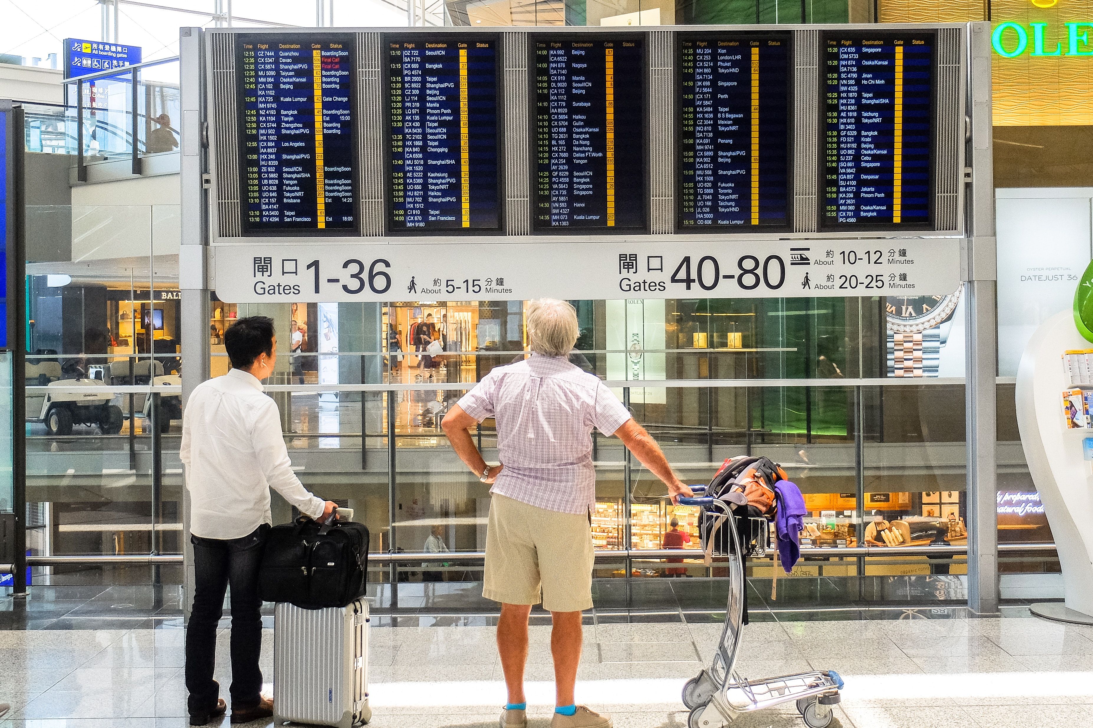 Two passengers look at screens in an airport terminal displaying upcoming arrivals and departures.