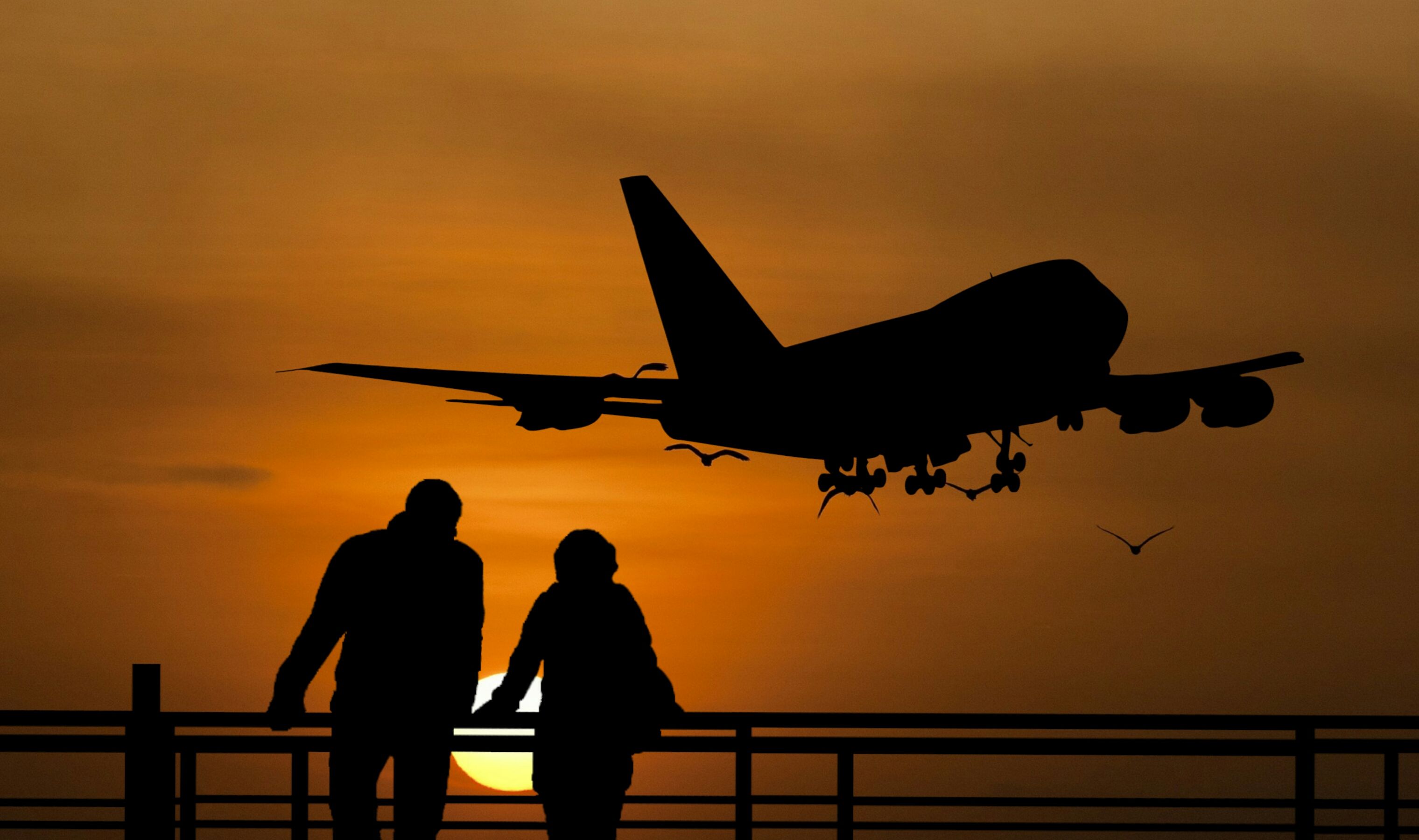 Two people look over a railing, during sunset, at a Boeing 747 taking off.
