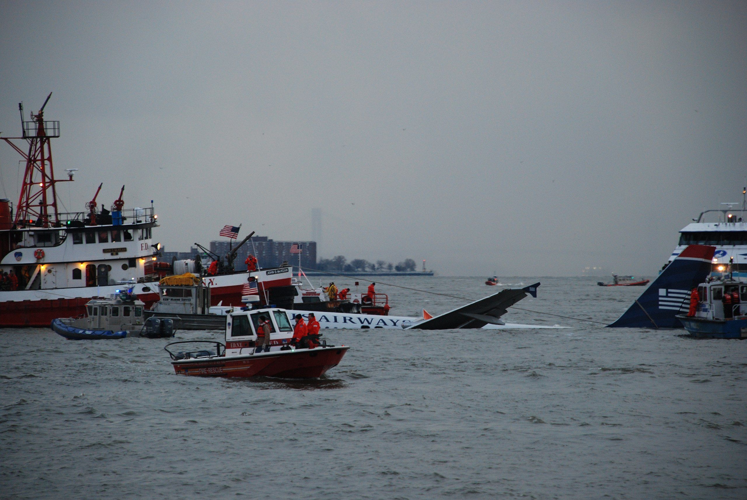 The top of the body and a portion of the wing of the US Airways A320 sticking out from the surface of the Hudson River surrounded by Coast Guard rescue boats.