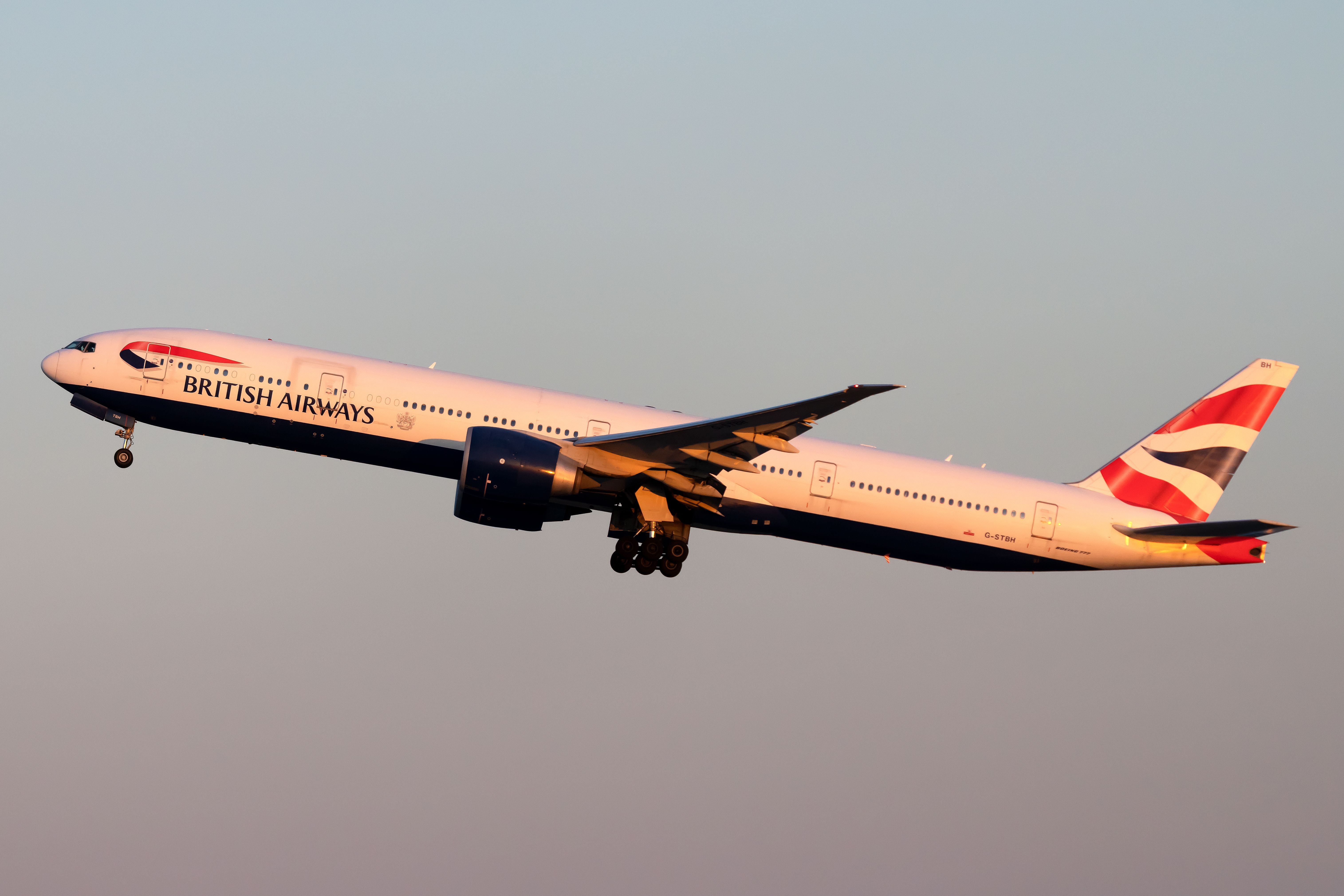 A British Airways Boeing 777-300 flying. In the background a slightly pink sky can be seen. 