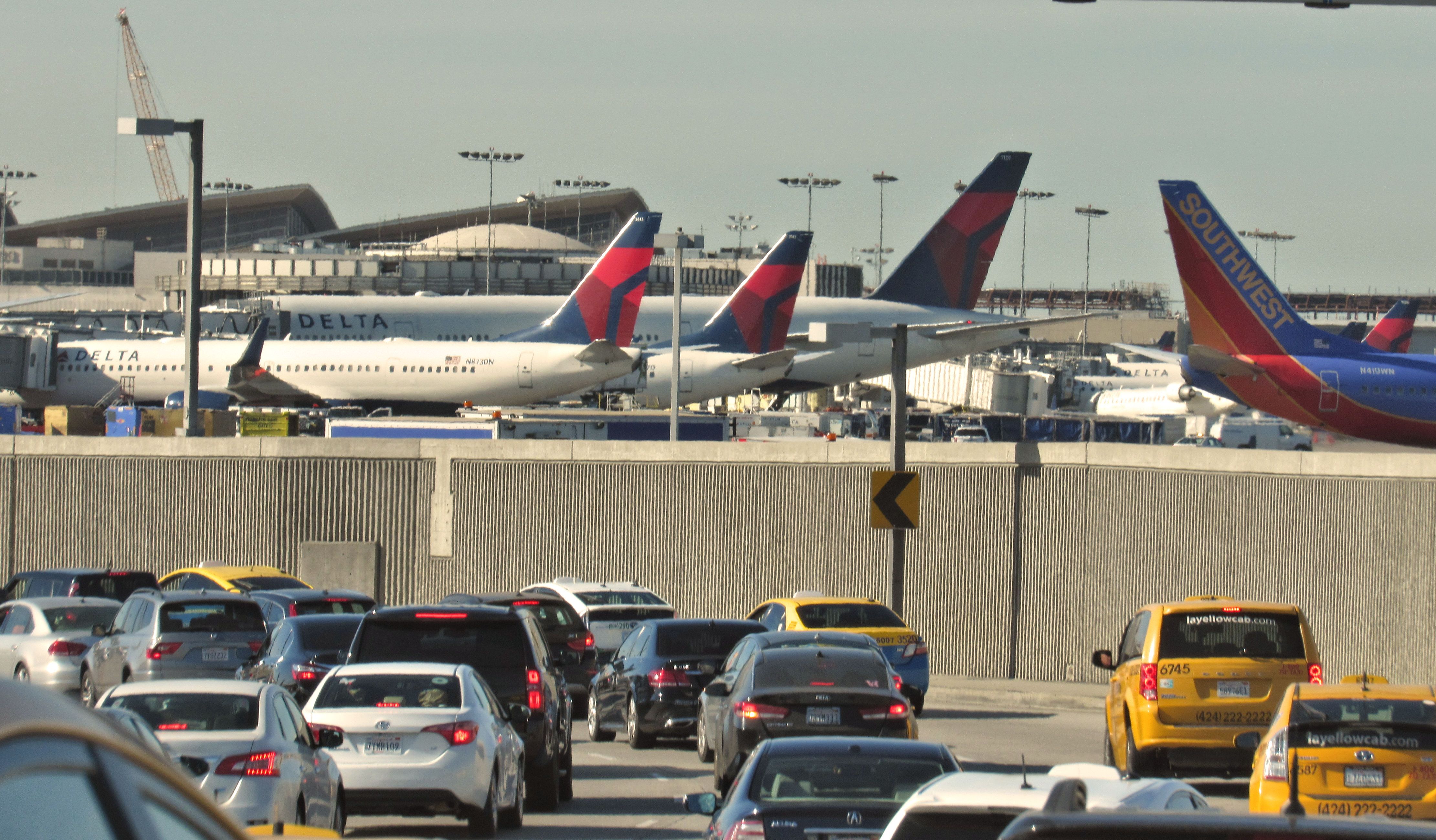 Road traffic files into LAX. Aircraft operated by Delta and Southwest are visible beyond the tarmac perimeter wall.