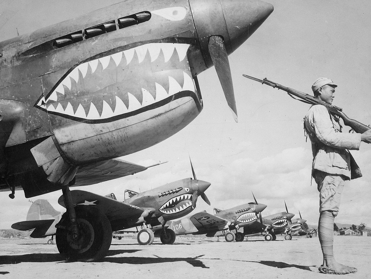 Several Curtiss P-40s with the famous shark mouth nose art parked on an airfield apron side by side.