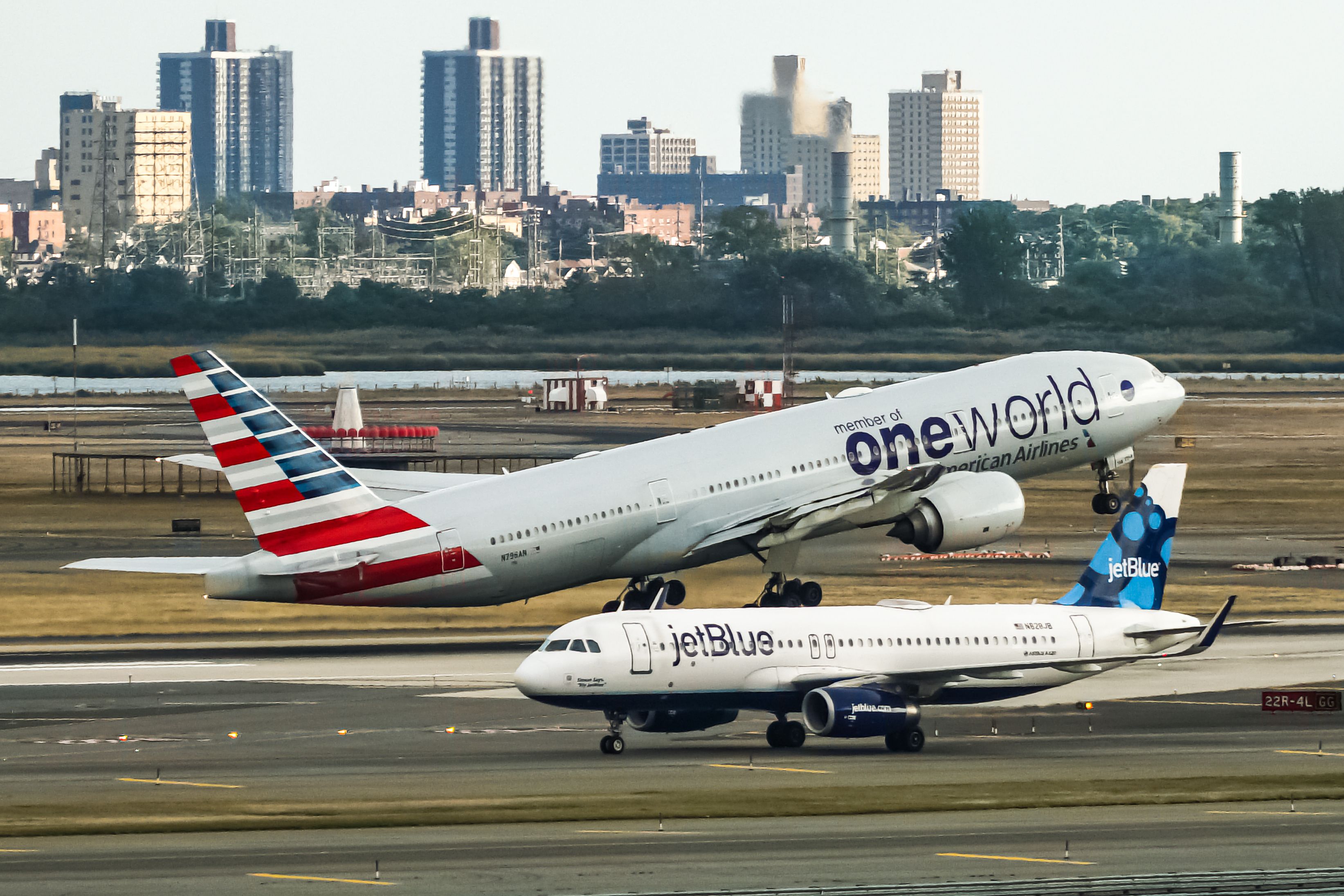 An American Airlines Boeing 777 in oneworld livery takes off while a JetBlue Airbus A320 taxis to the runway at JFK airport.