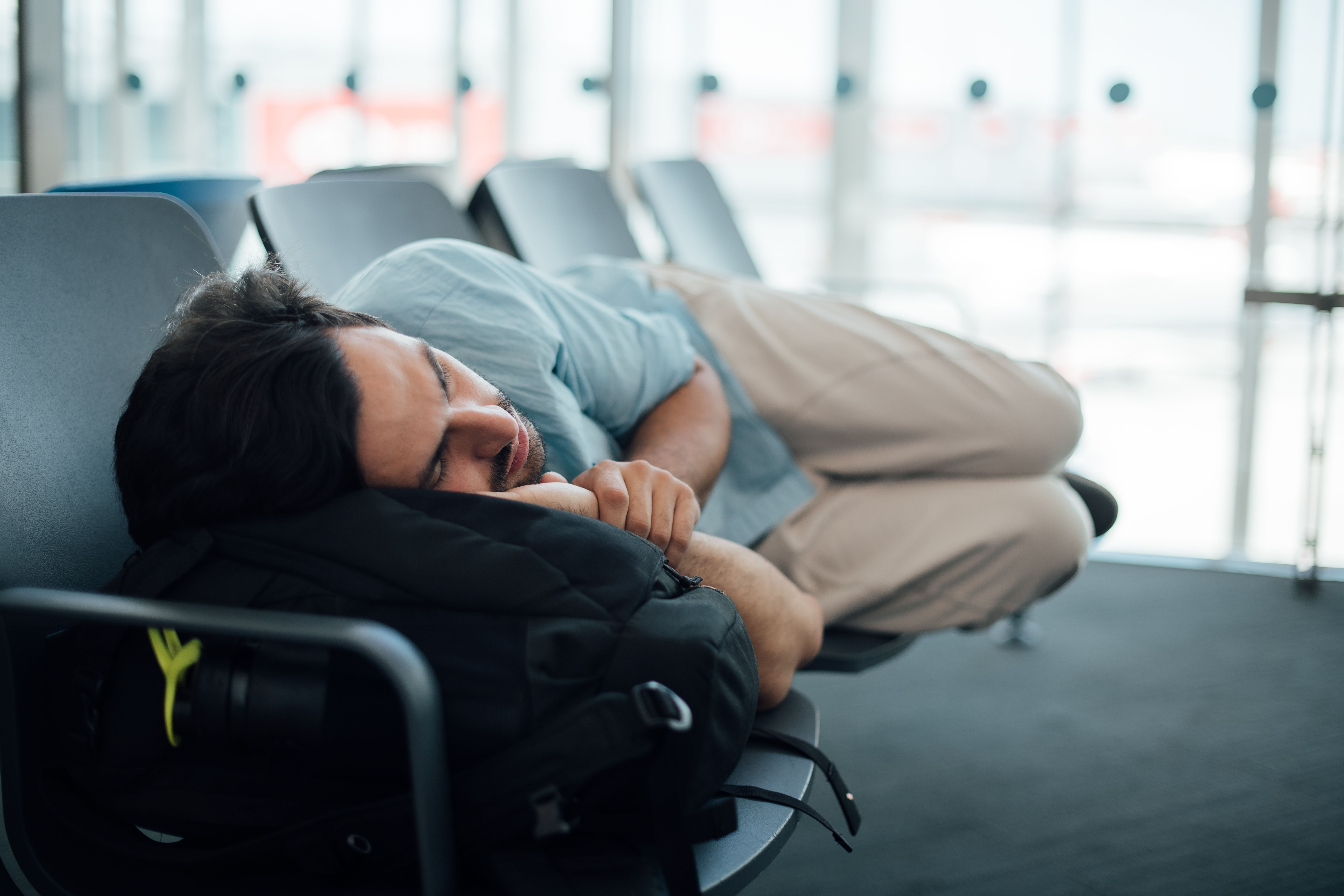 A man sleeping on a few airport gate seats, waiting to board an aircraft.