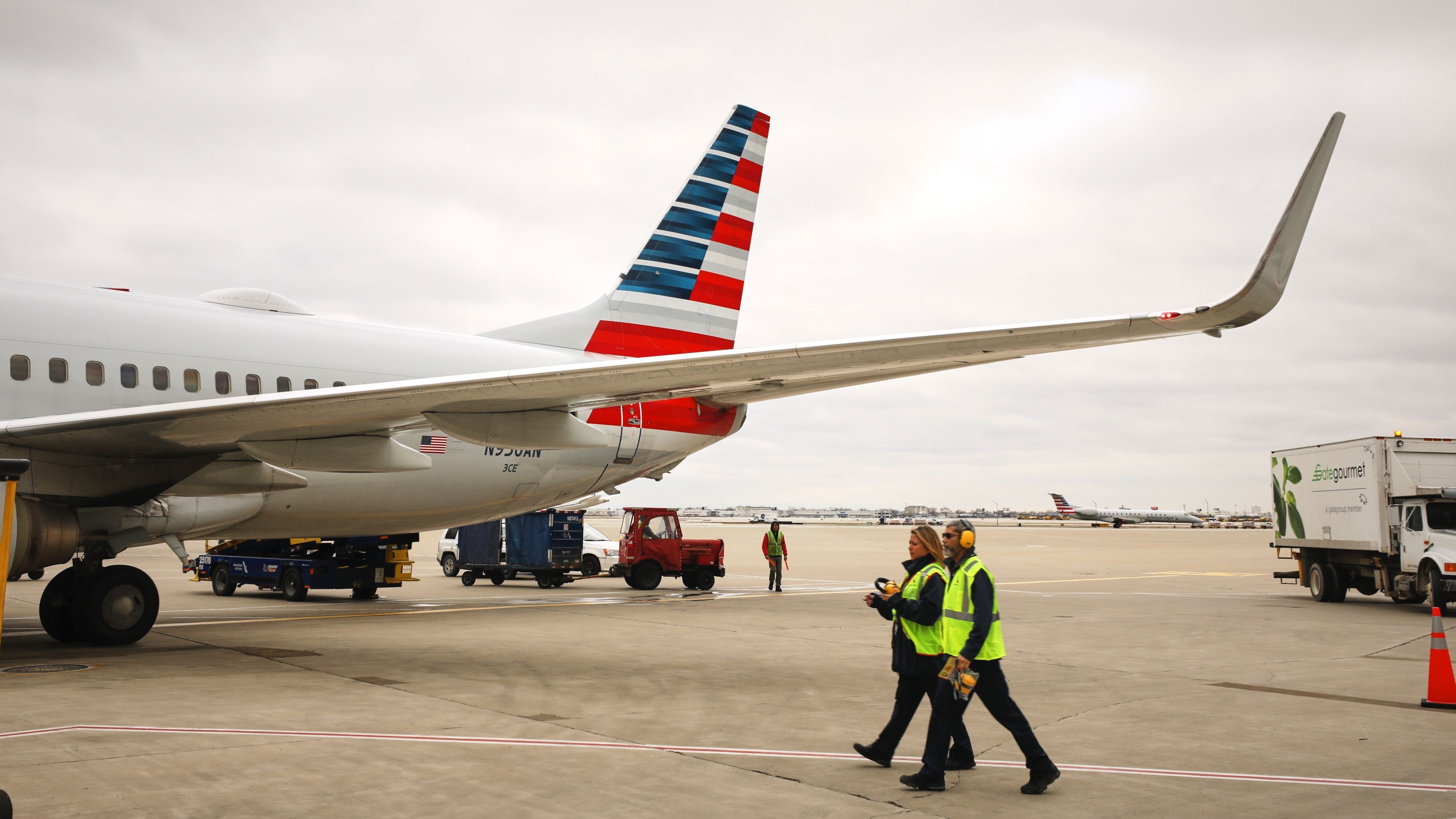 Two crew members in bright yellow safety vests walk toward the mid-section of a parked American Airlines aircraft.