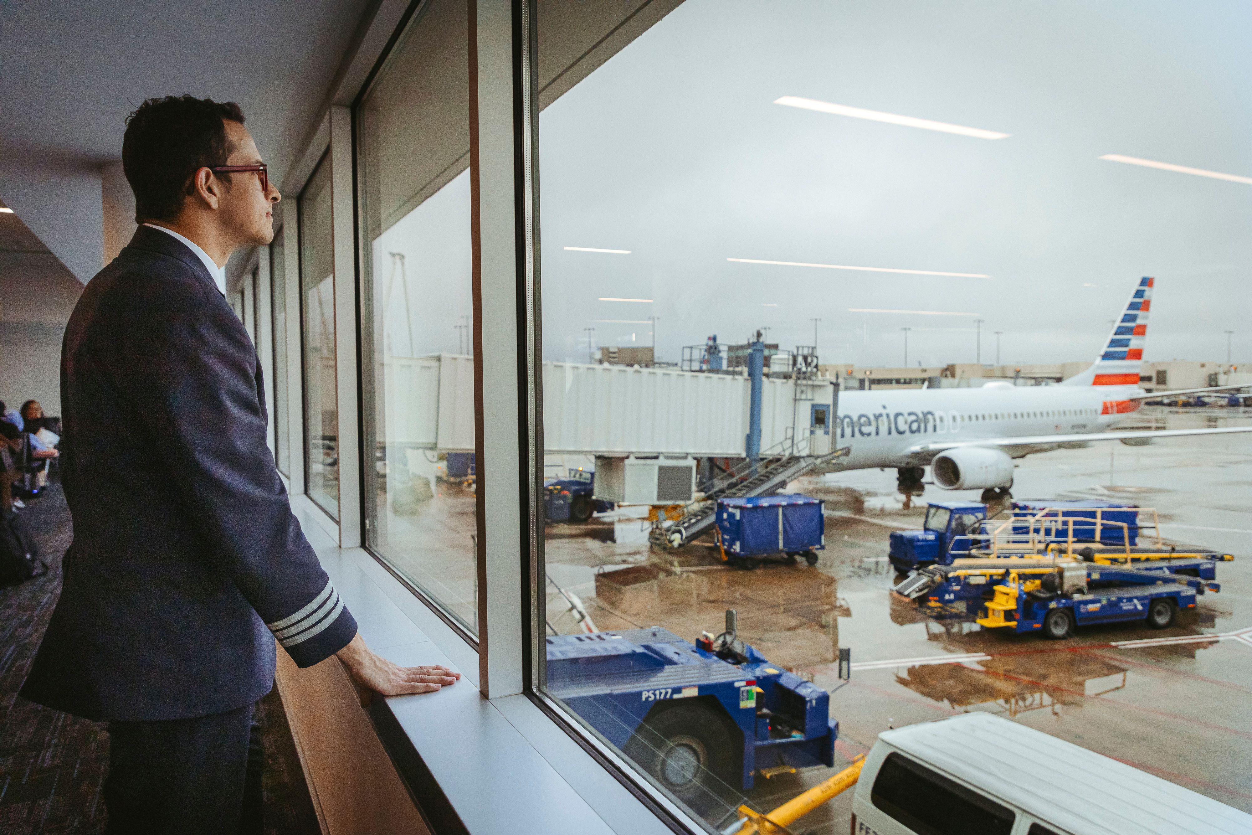 Man in a navy blue pilot's uniform looks out the window at American Airlines passenger jets parked at gates