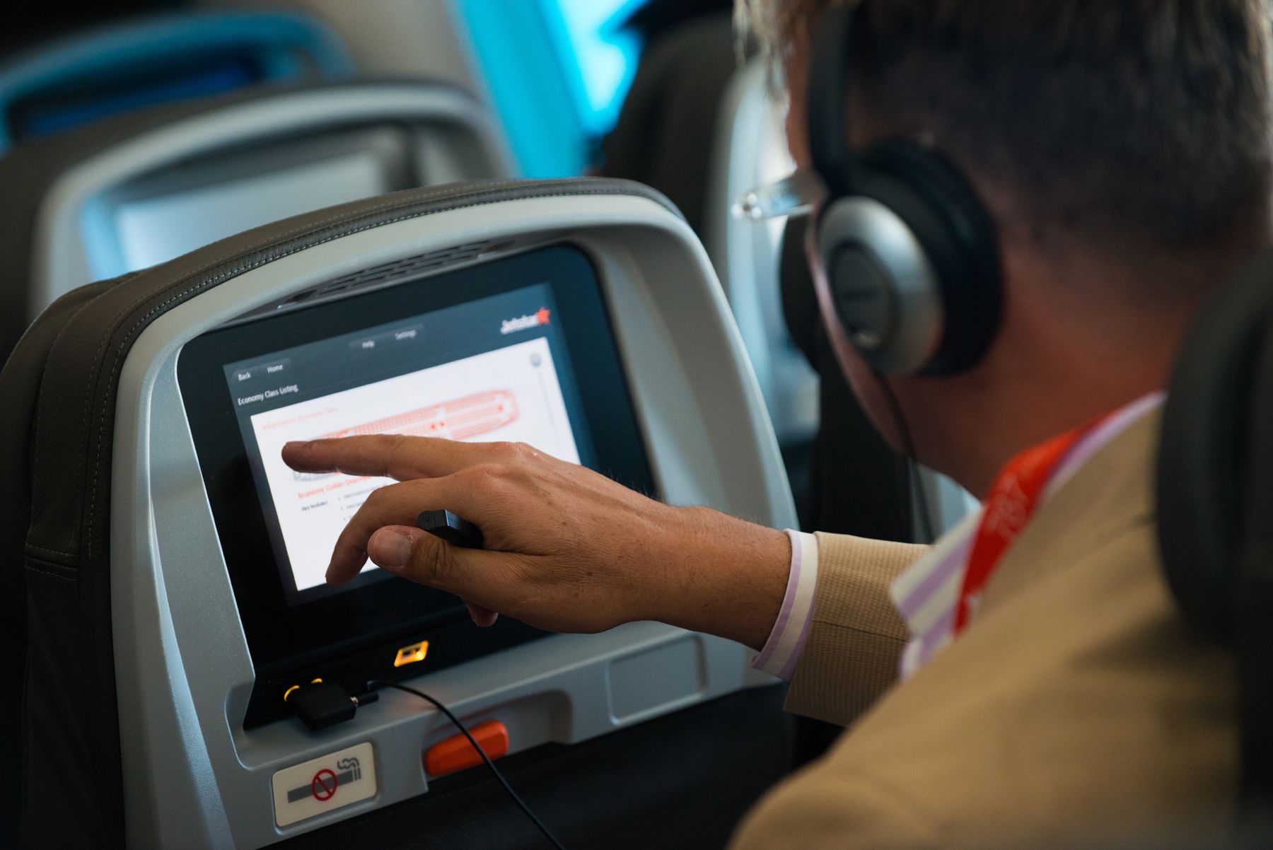 Man in a beige jacket using the touch screen in a seatback entertainment system wearing headphones