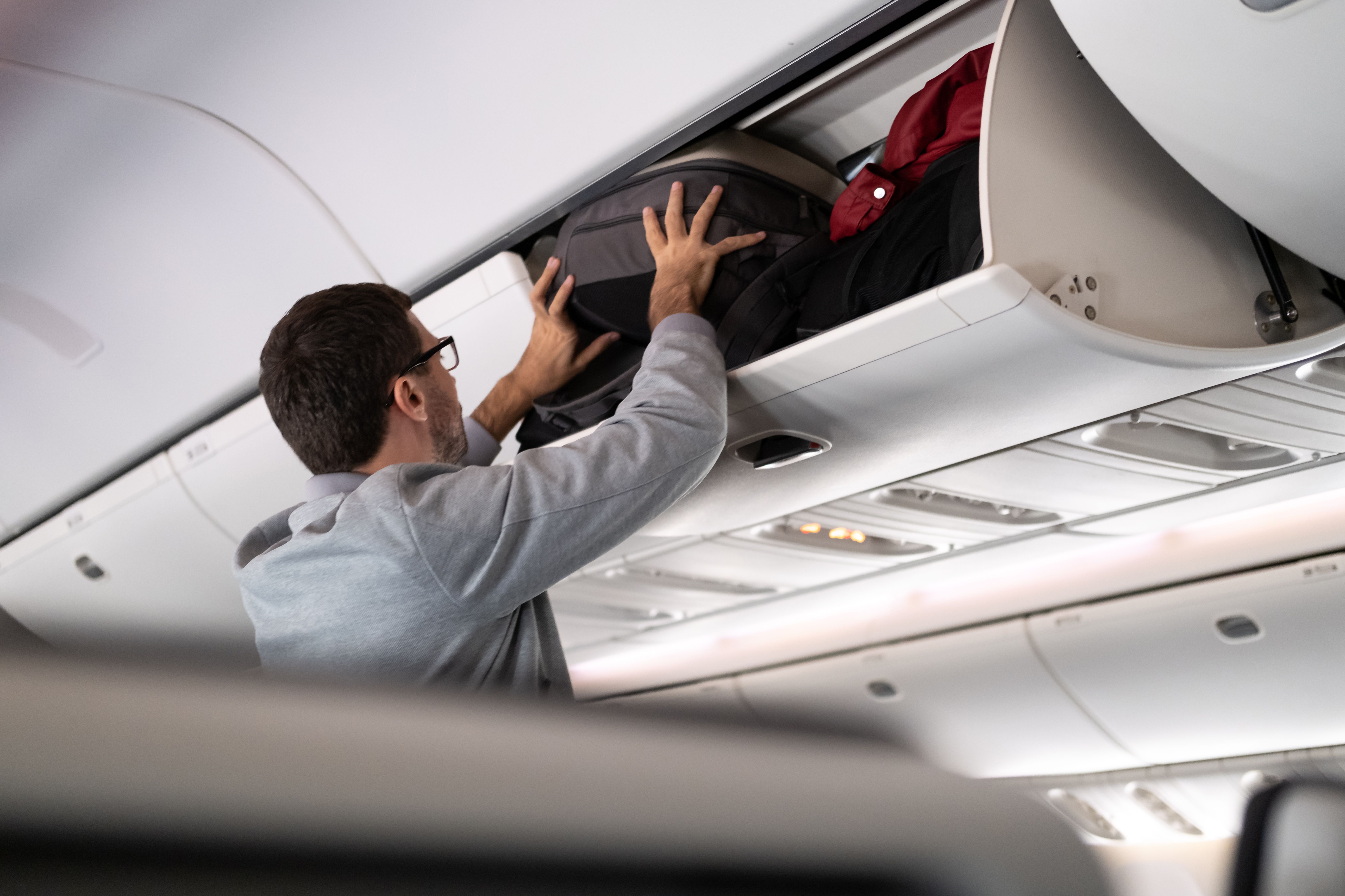 A man forces a bag into a full overhead bin on an aircraft.