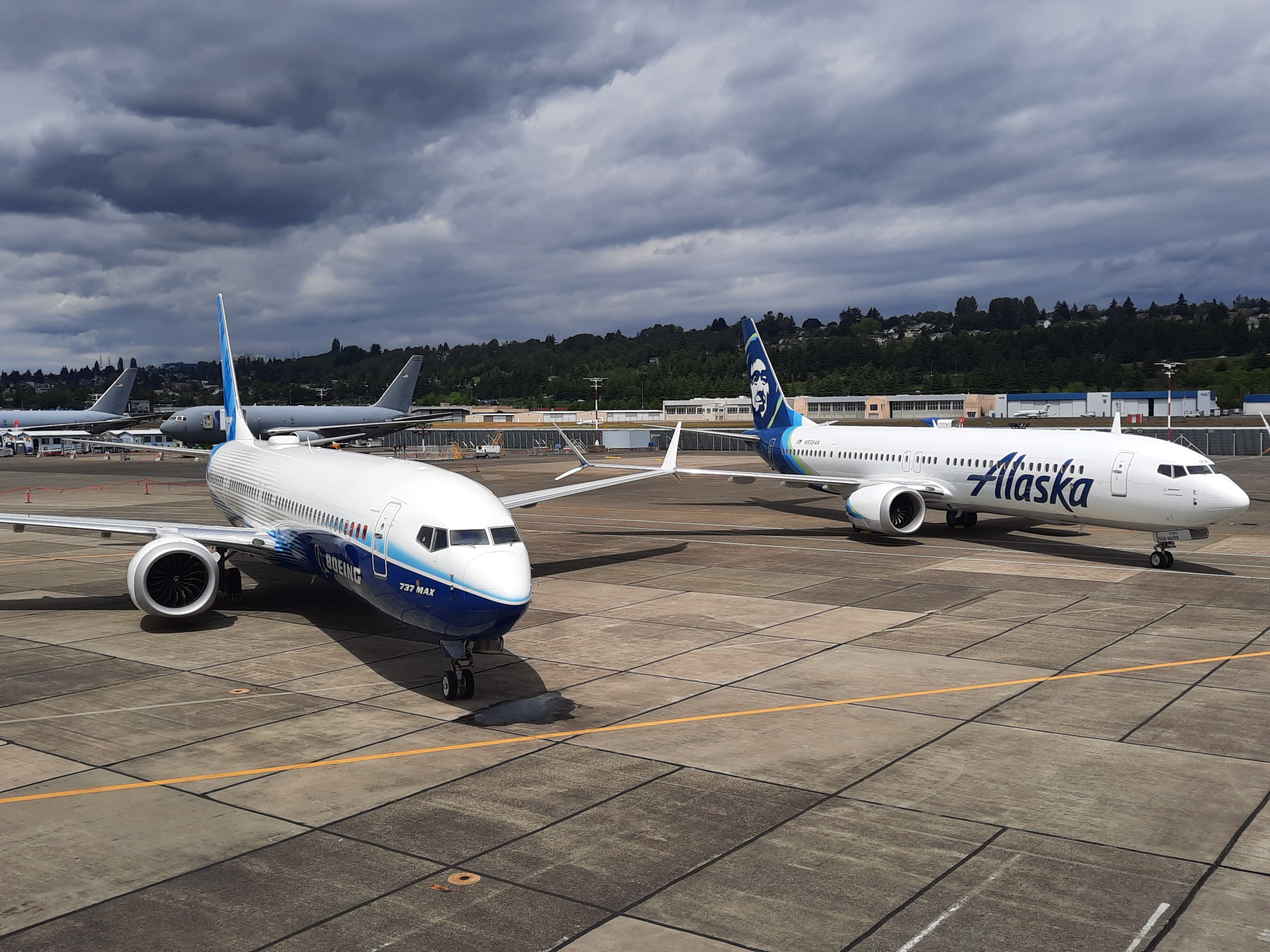 Two Boeing 737 MAX Aircrat Parked In Sunny Conditions With Military Planes Behind Them.