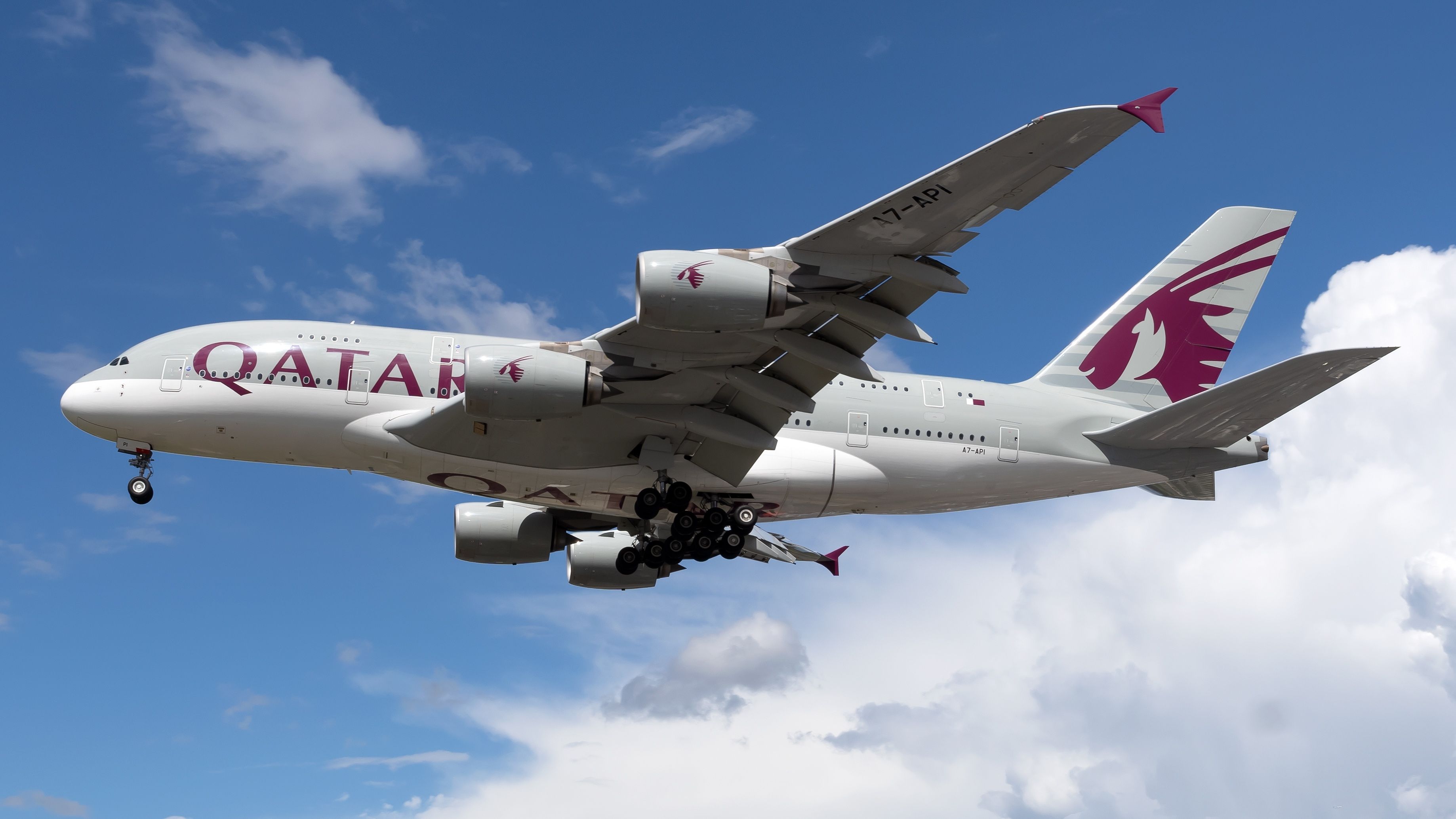 White Airbus A380 jetliner with Qatar in purple lettering on the side flying through blue sky and clouds