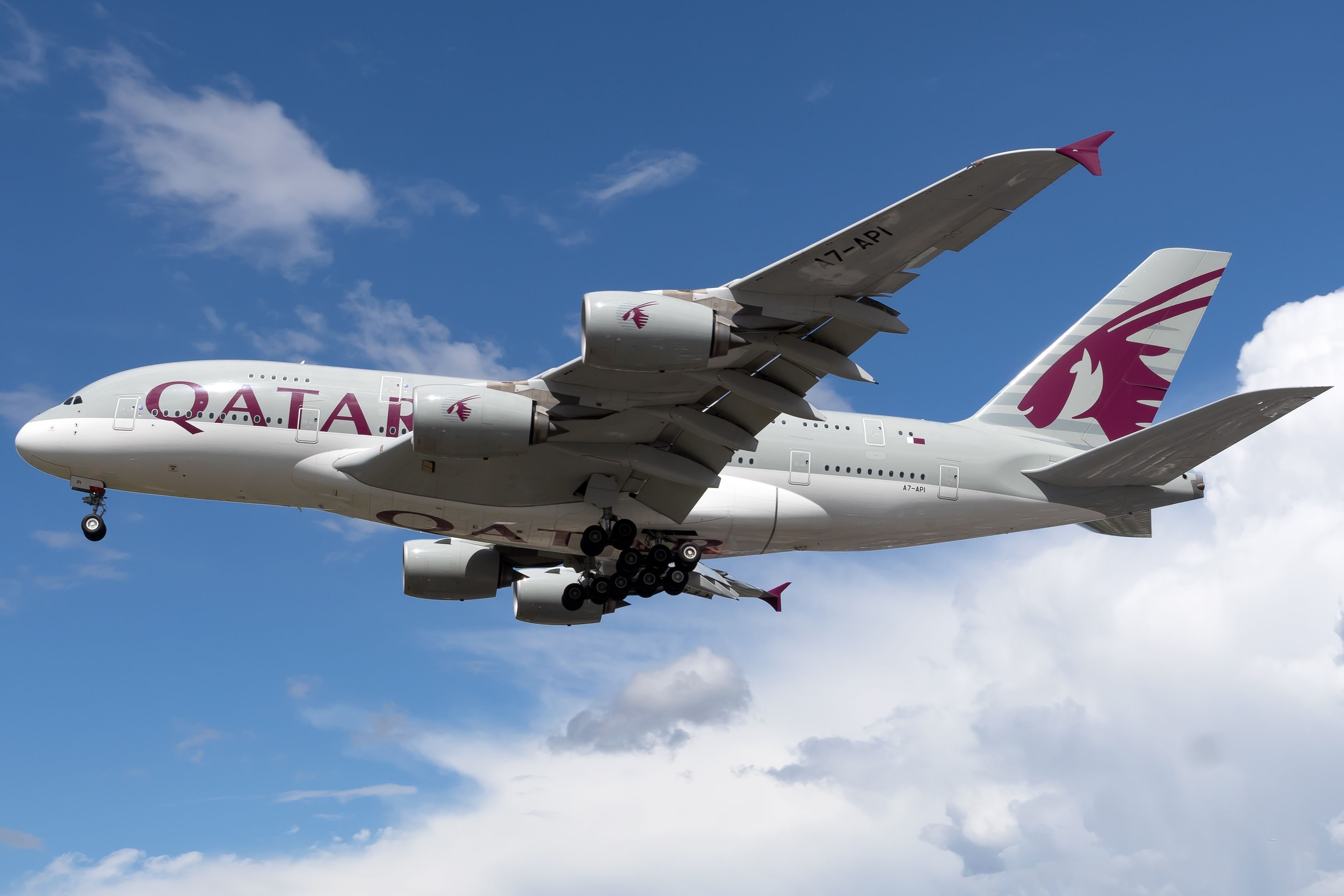 White Airbus A380 jetliner with Qatar in purple lettering on the side flying in blue sky and clouds