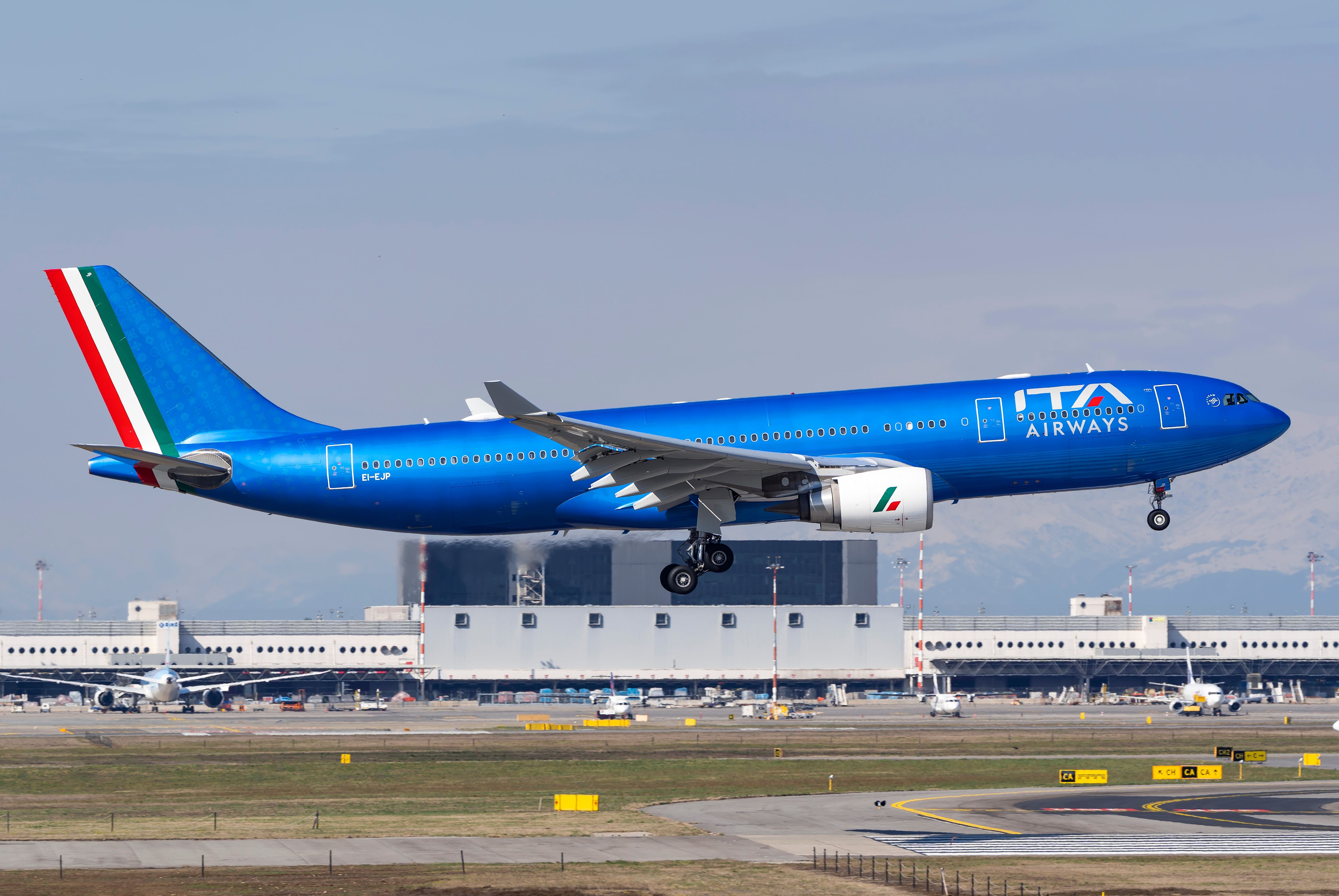 An ITA Airways Airbus A330-200 taking off with an airport in the background.