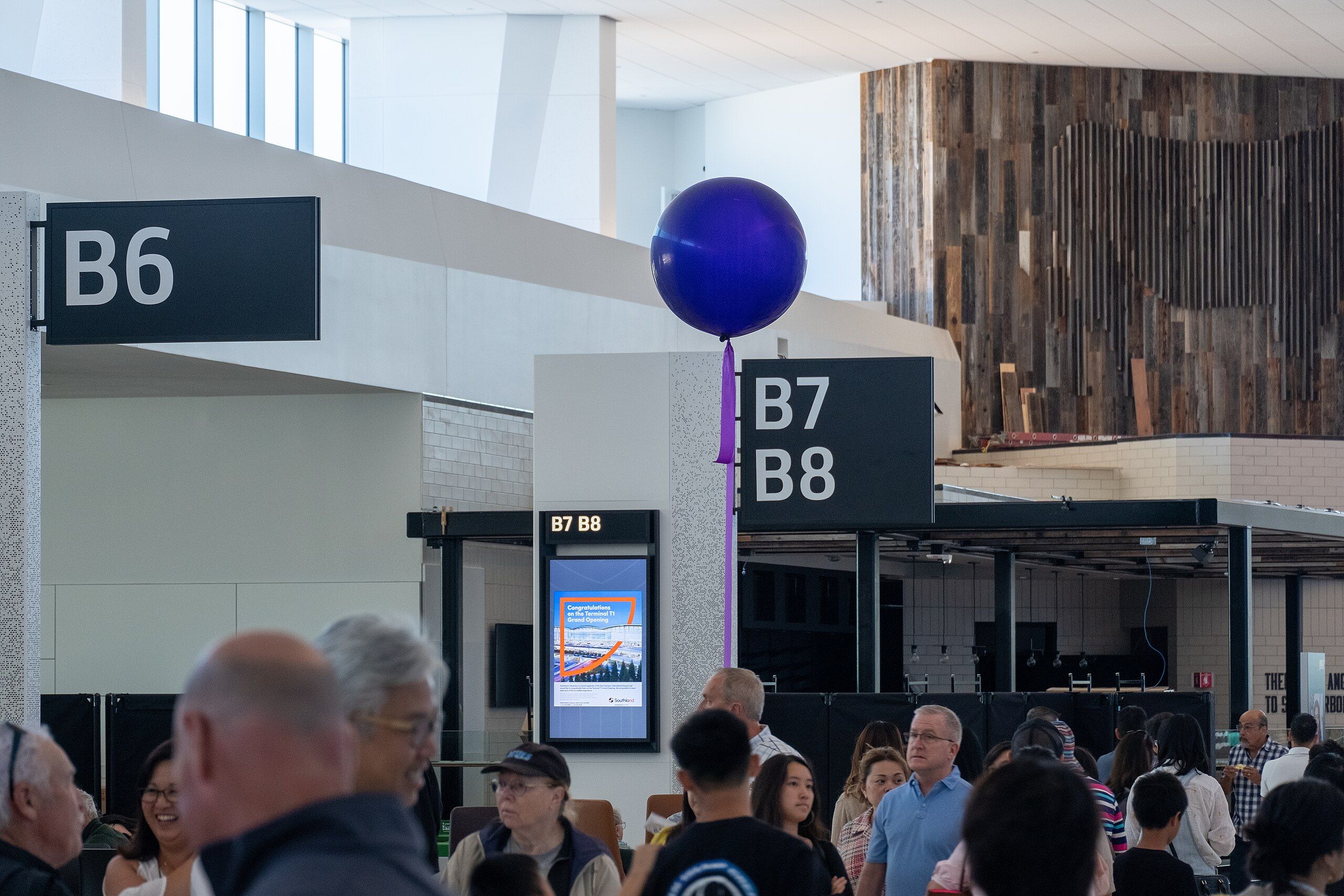 Passengers gathered underneath signs marking B gates with sunlight coming in through tall windows