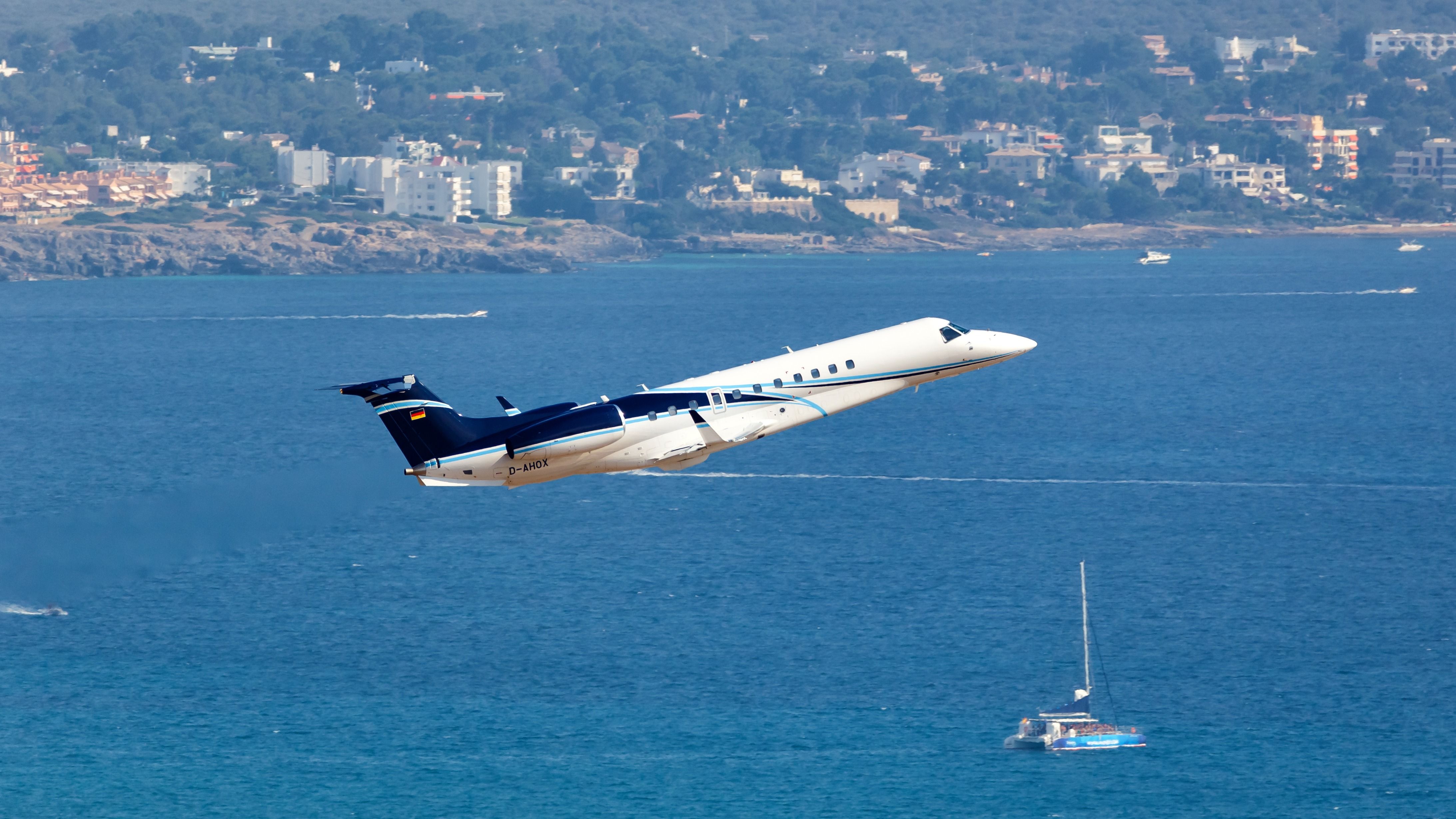 An Embraer Legacy 650 private jet taking off with a yacht-filled ocean in the background.