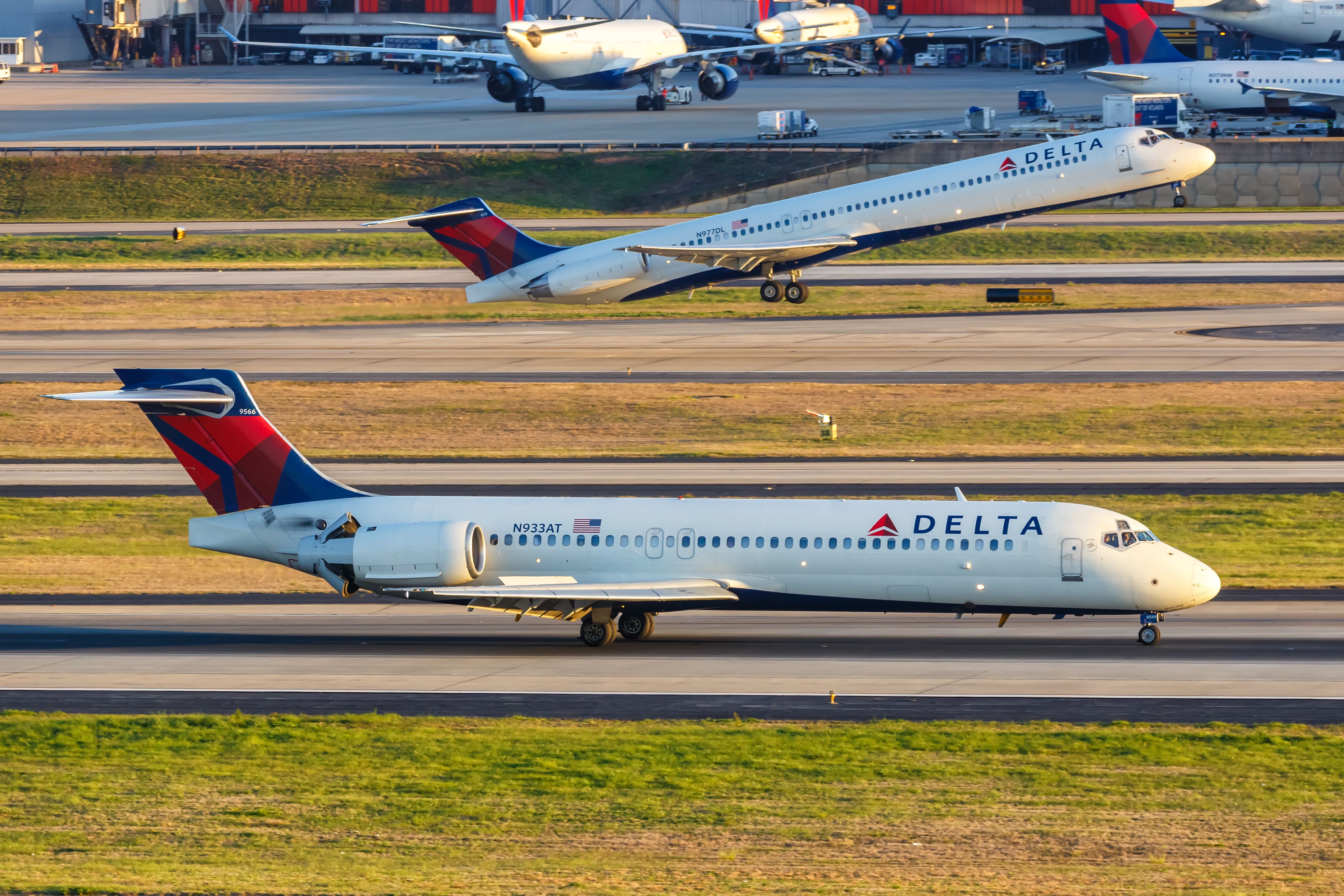 A Delta Air Lines Boeing 717 on a taxiway, as another Delta Air Lines aircraft takes off in the background.