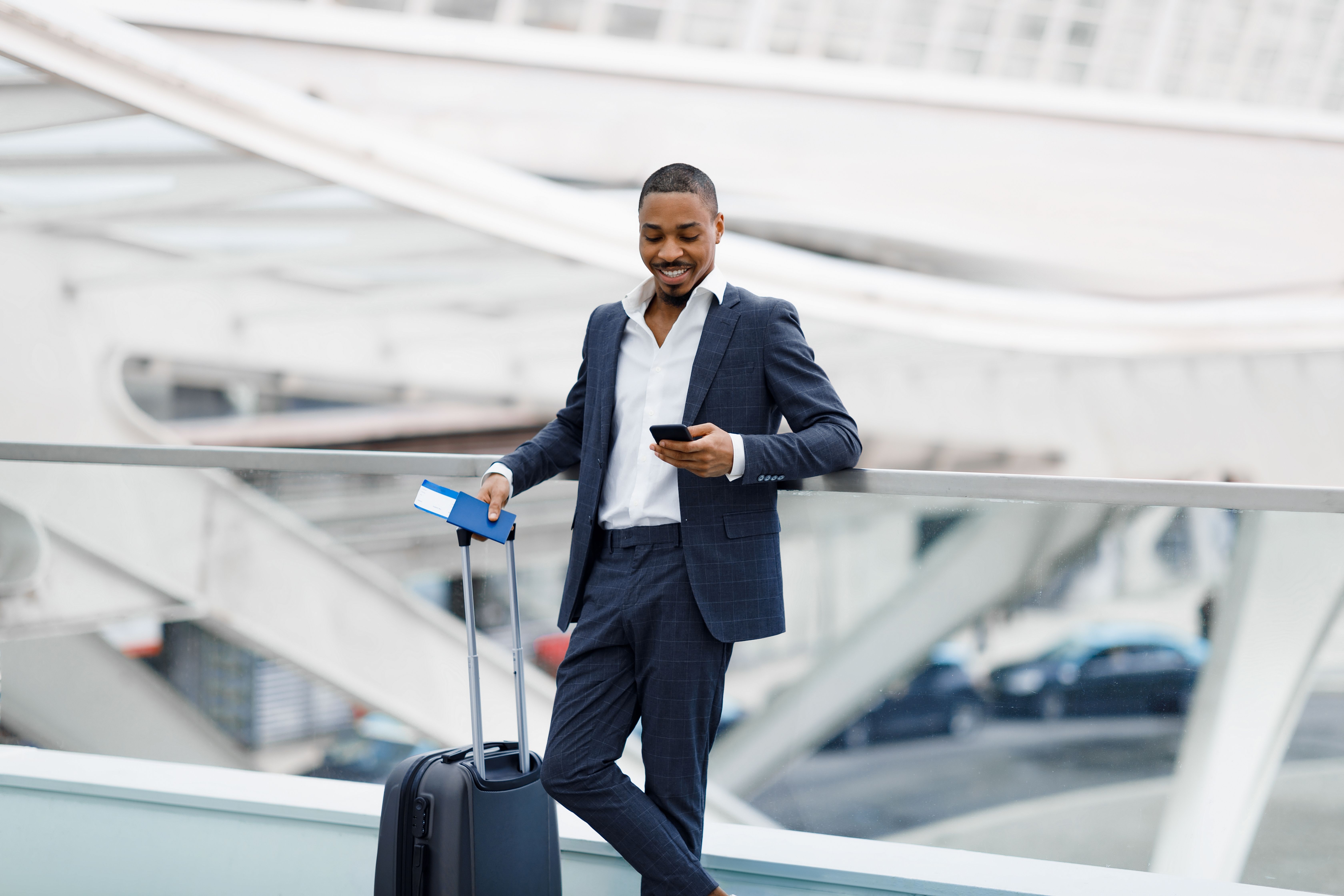 A man in a suit using a mobile phone standing next to a small suitcase and carrying a passport and boarding pass.