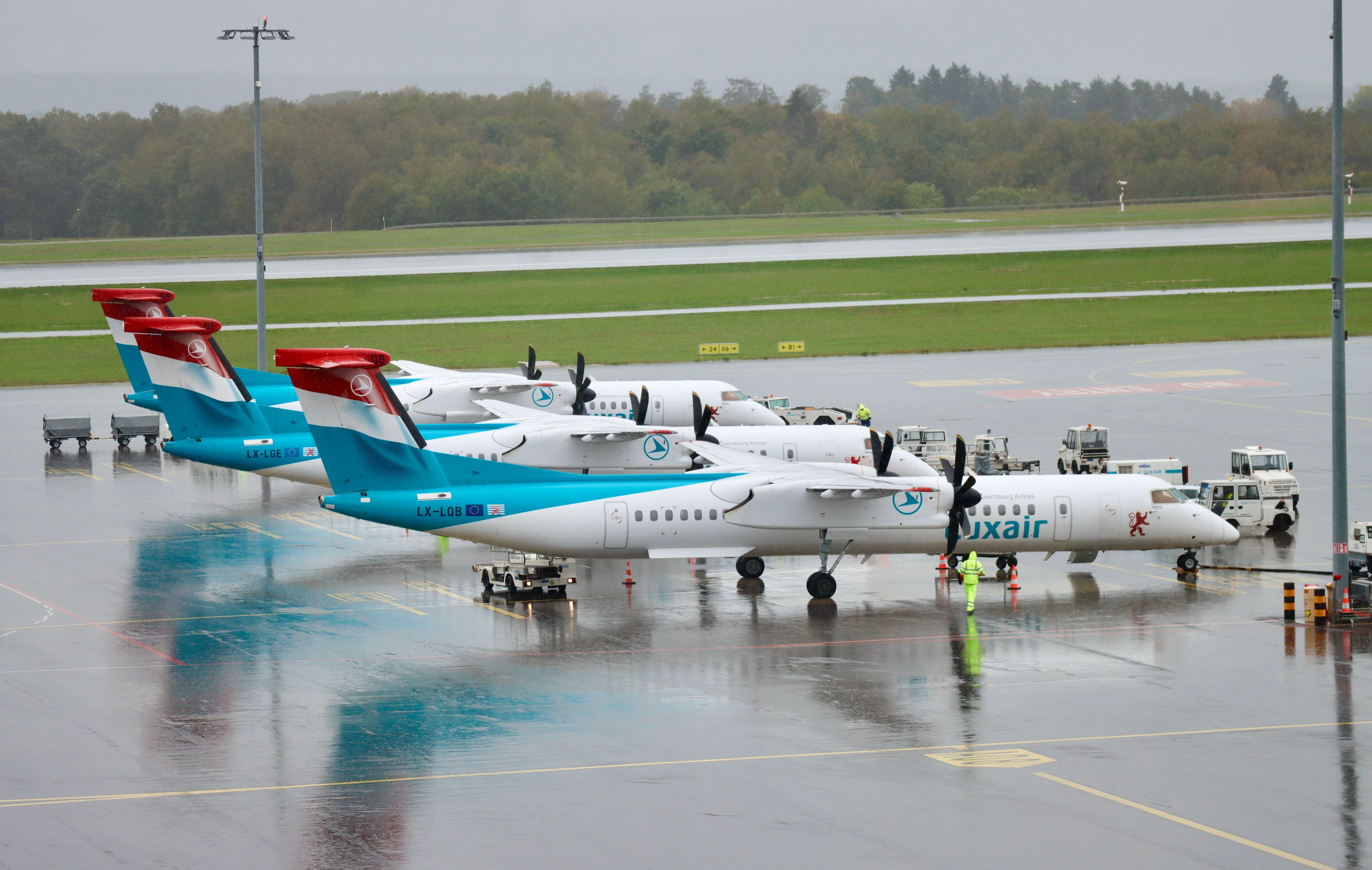 Multiuple Luxair Dash 8s Lined Up on a wet airport apron.