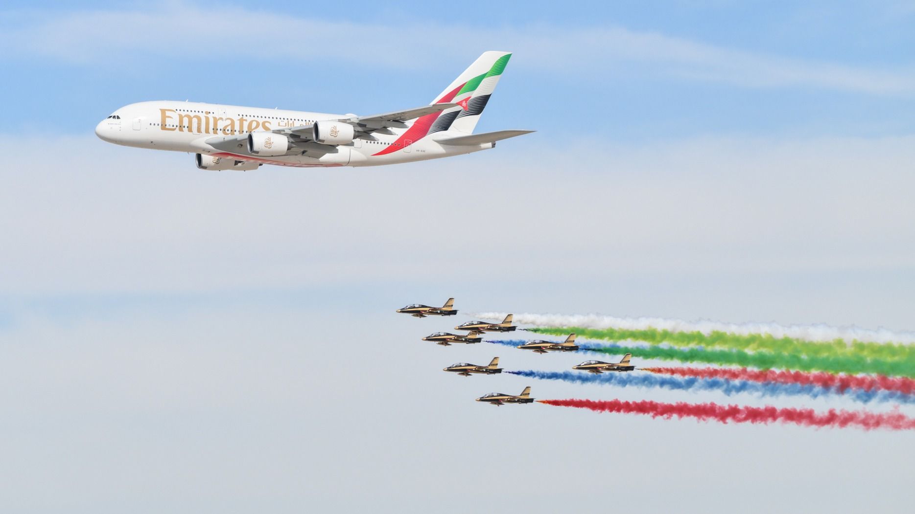 An Airbus A380 of the Emirates airline is pursued by several fighter jets that emit colored smoke behind them.