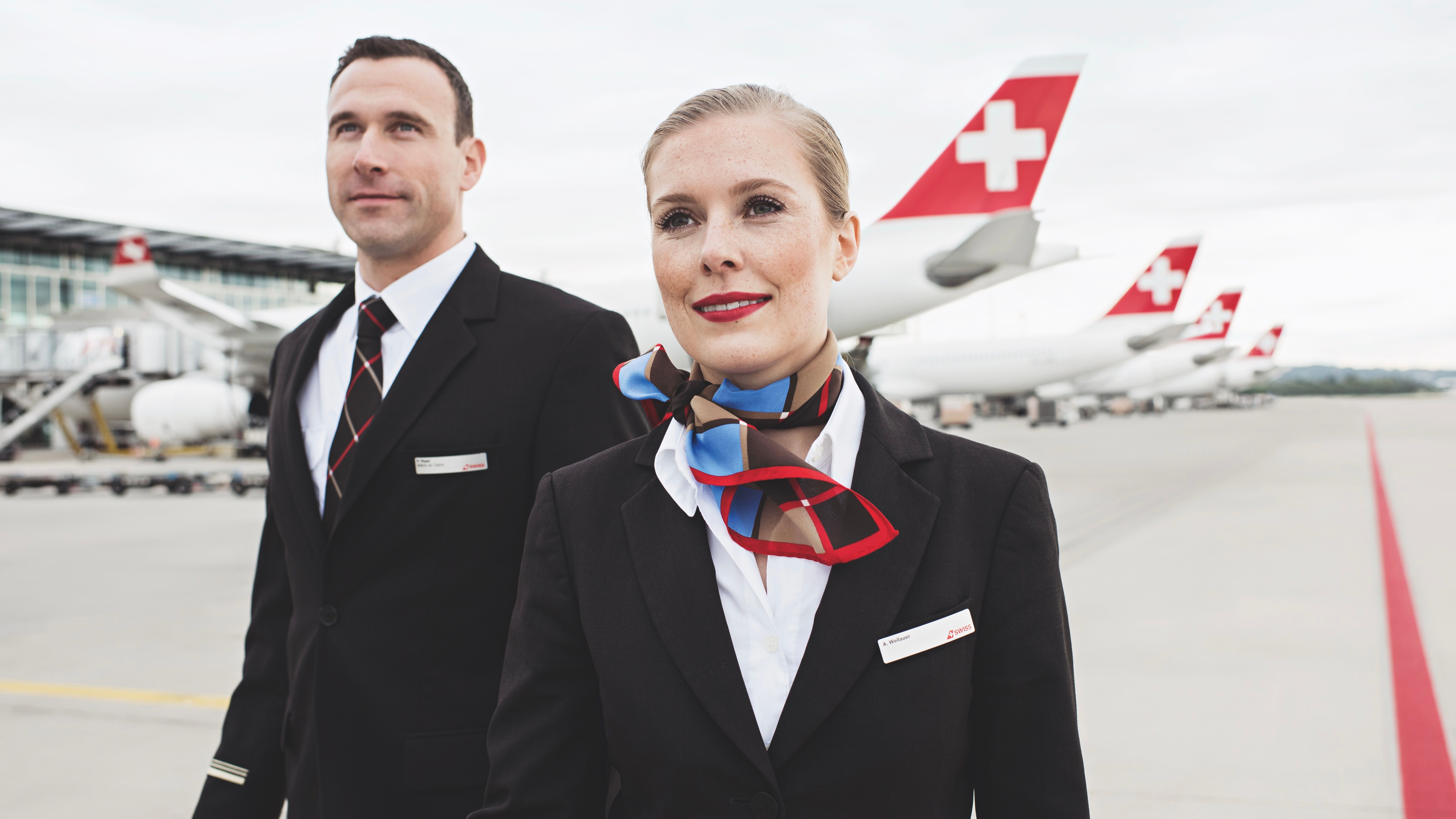 Two SWISS cabin crew members standing on the airport apron in front of several large aircraft.