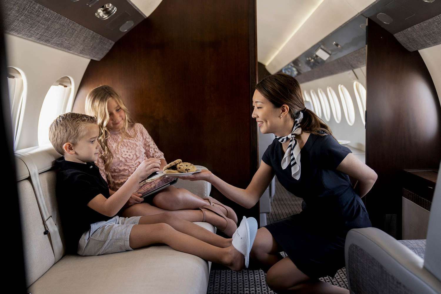 A flight attendant offers cookies to two young passengers on a private jet.