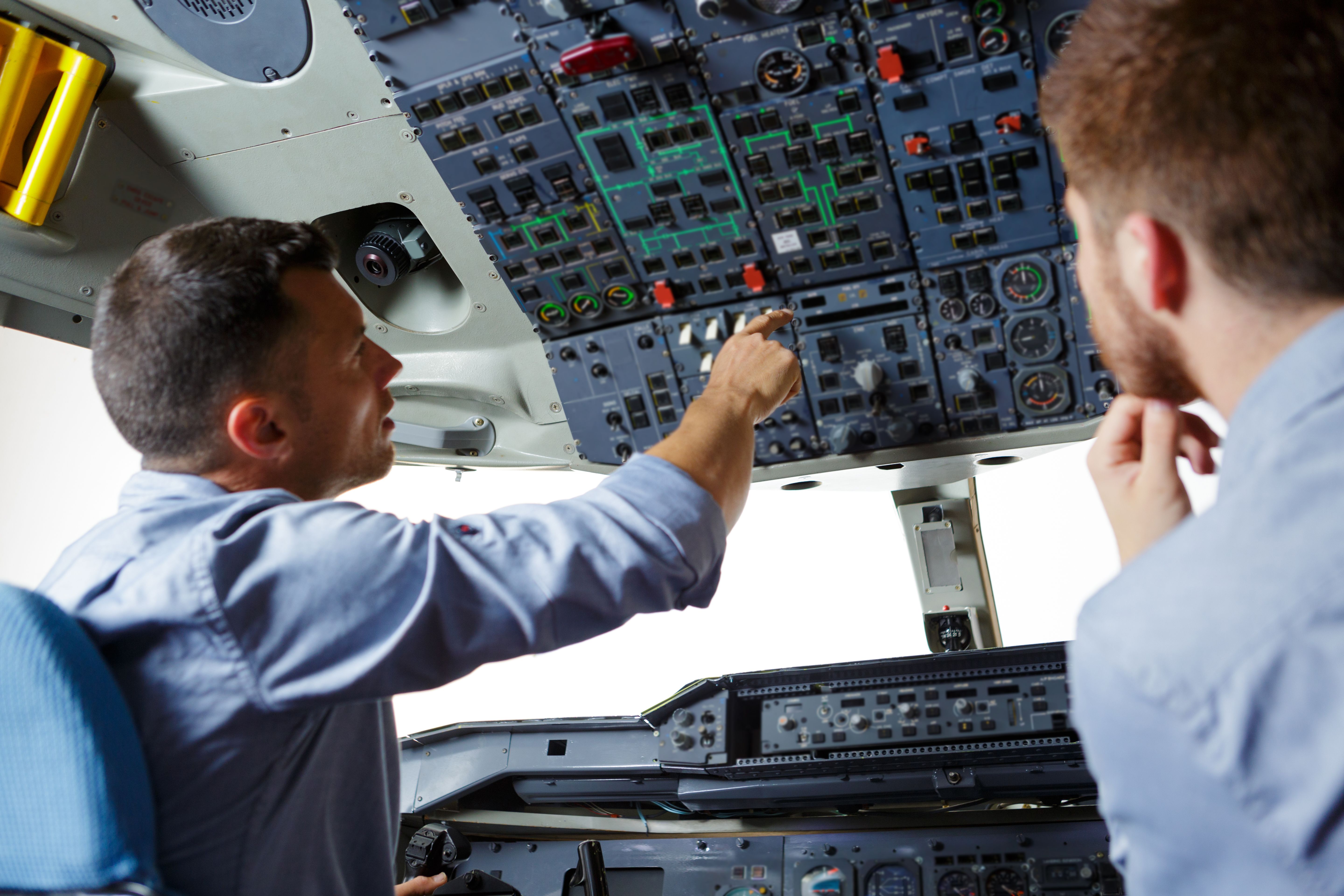 Two pilots inspecting buttons on the overhead panel of an aircraft cockpit.