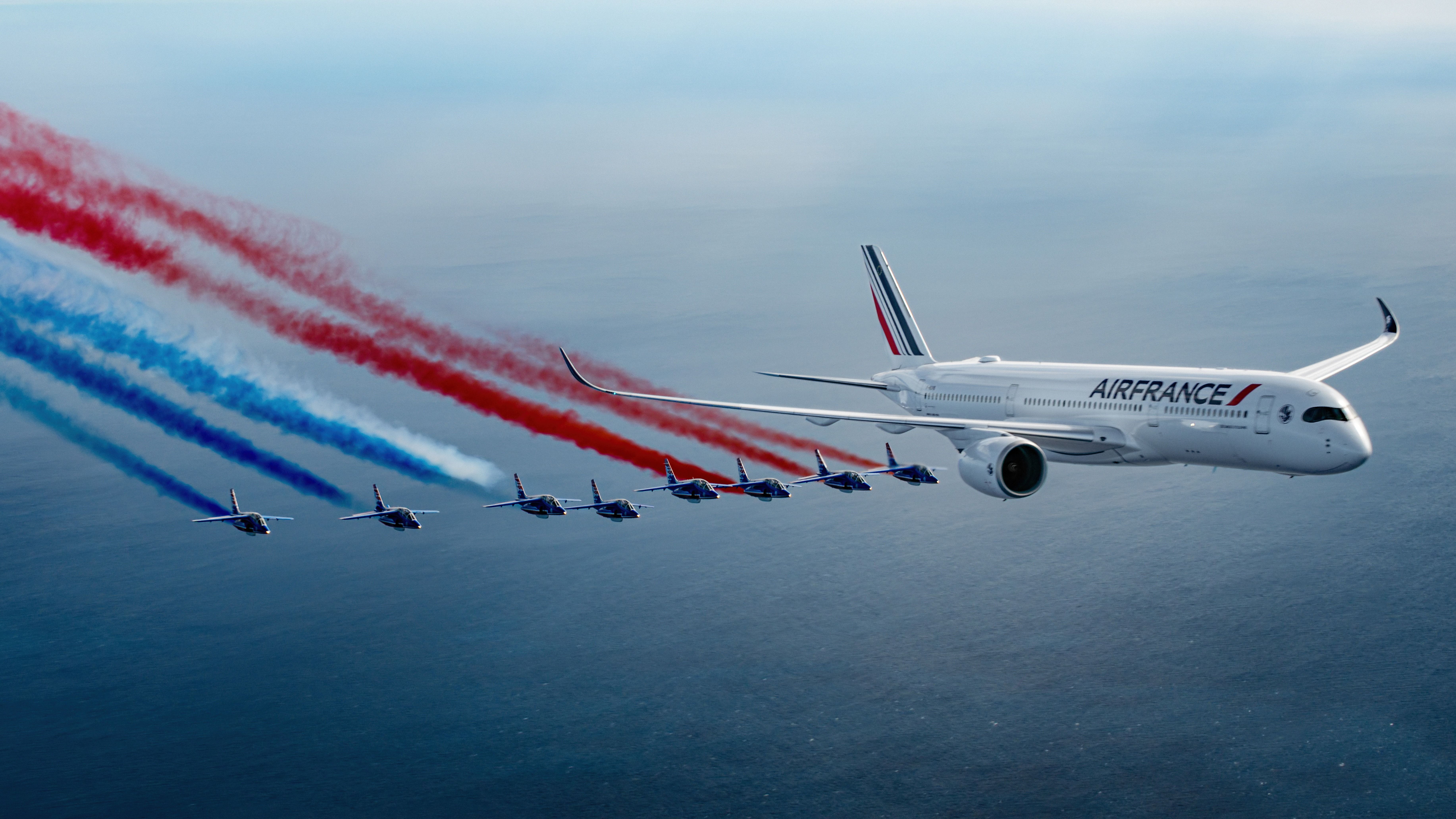 An Air France Airbus A350 flying in front of several fighter jets giving off blue, white, and red smoke.