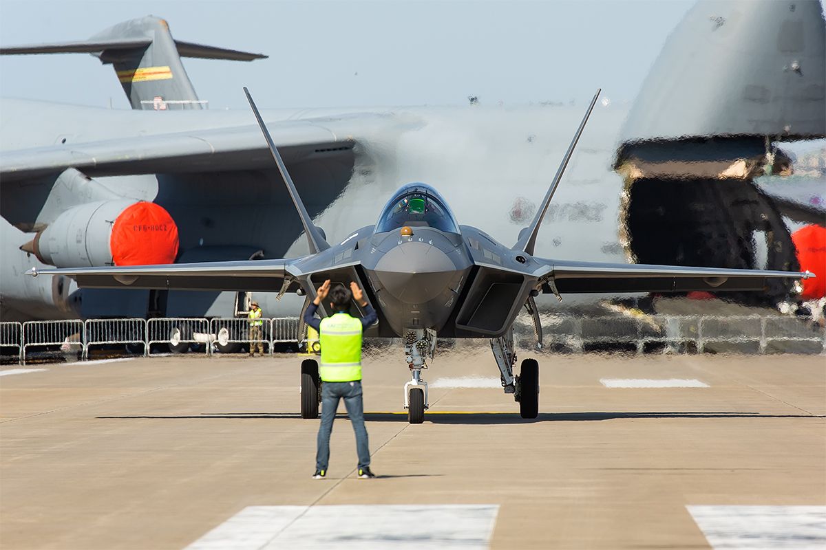 A KAI KF-21 on an airfield with a massive transport aircraft in the background.