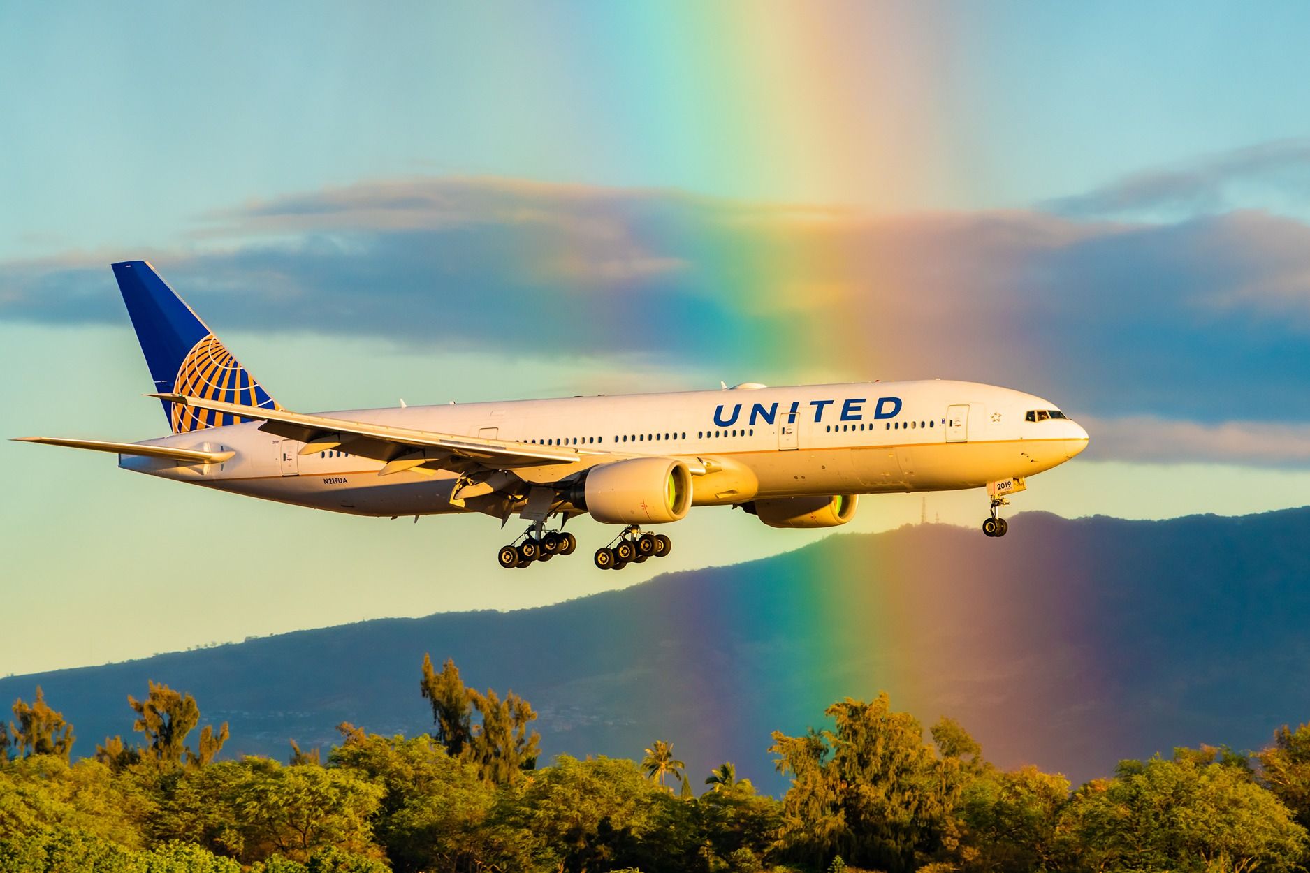 A United Airlines Boeing 777-200 about to land with mountains and a rainbow in the background.