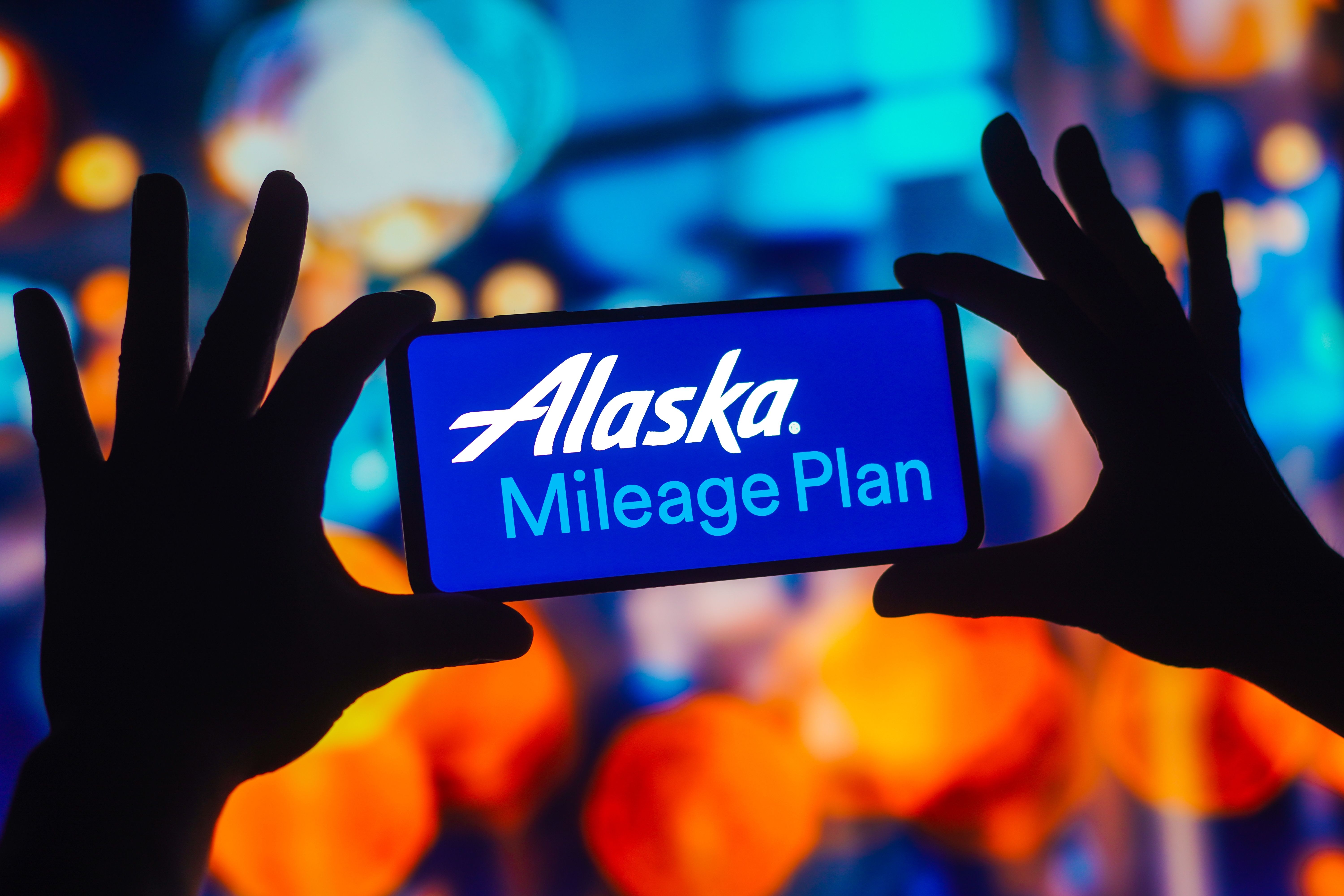 A person holding up a phone with the logo for Alaska Airlines Mileage Plan on it.