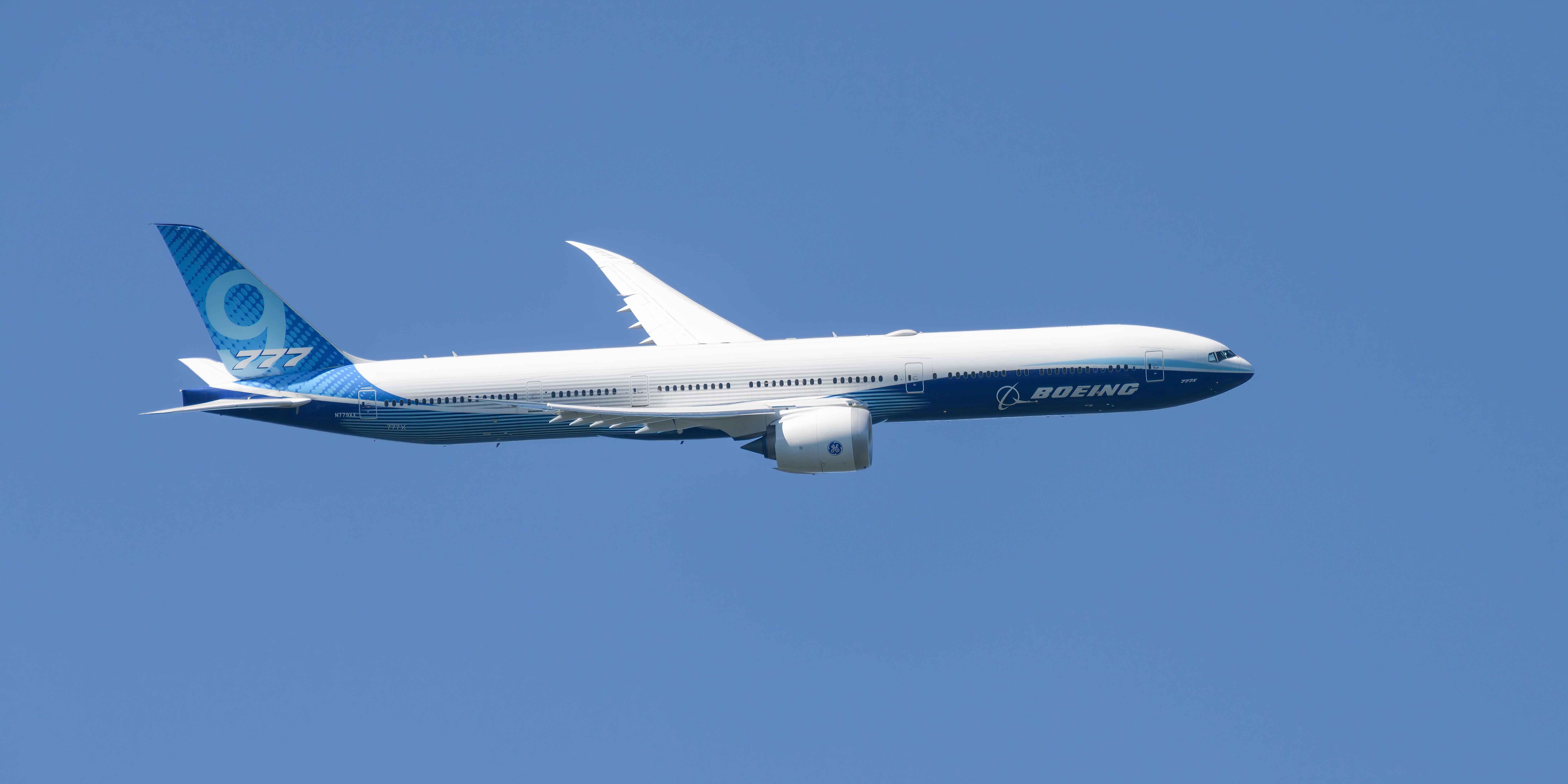 Side view of a new 777X in flight turning against a blue sky