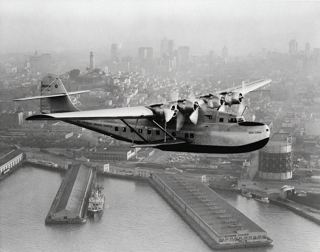 The Martin China Clipper flying over the water near a city. 